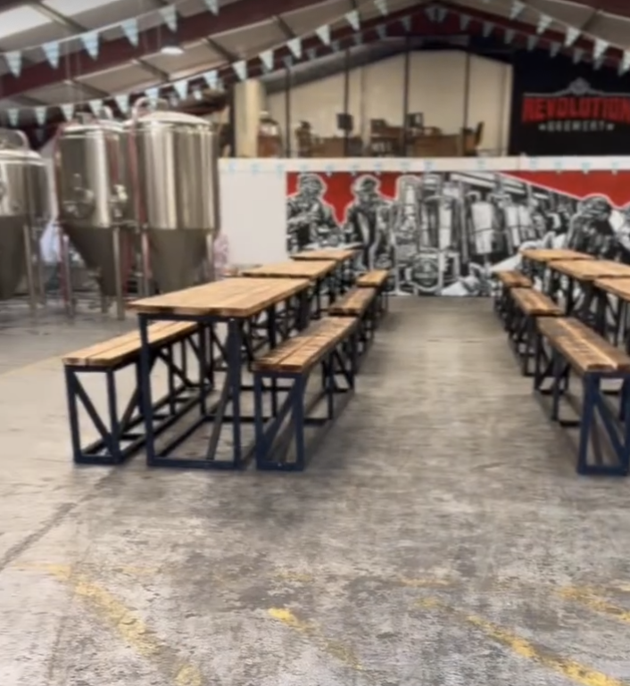 Empty picnic tables and brewery equipment in an industrial-style space with a mural of workers.