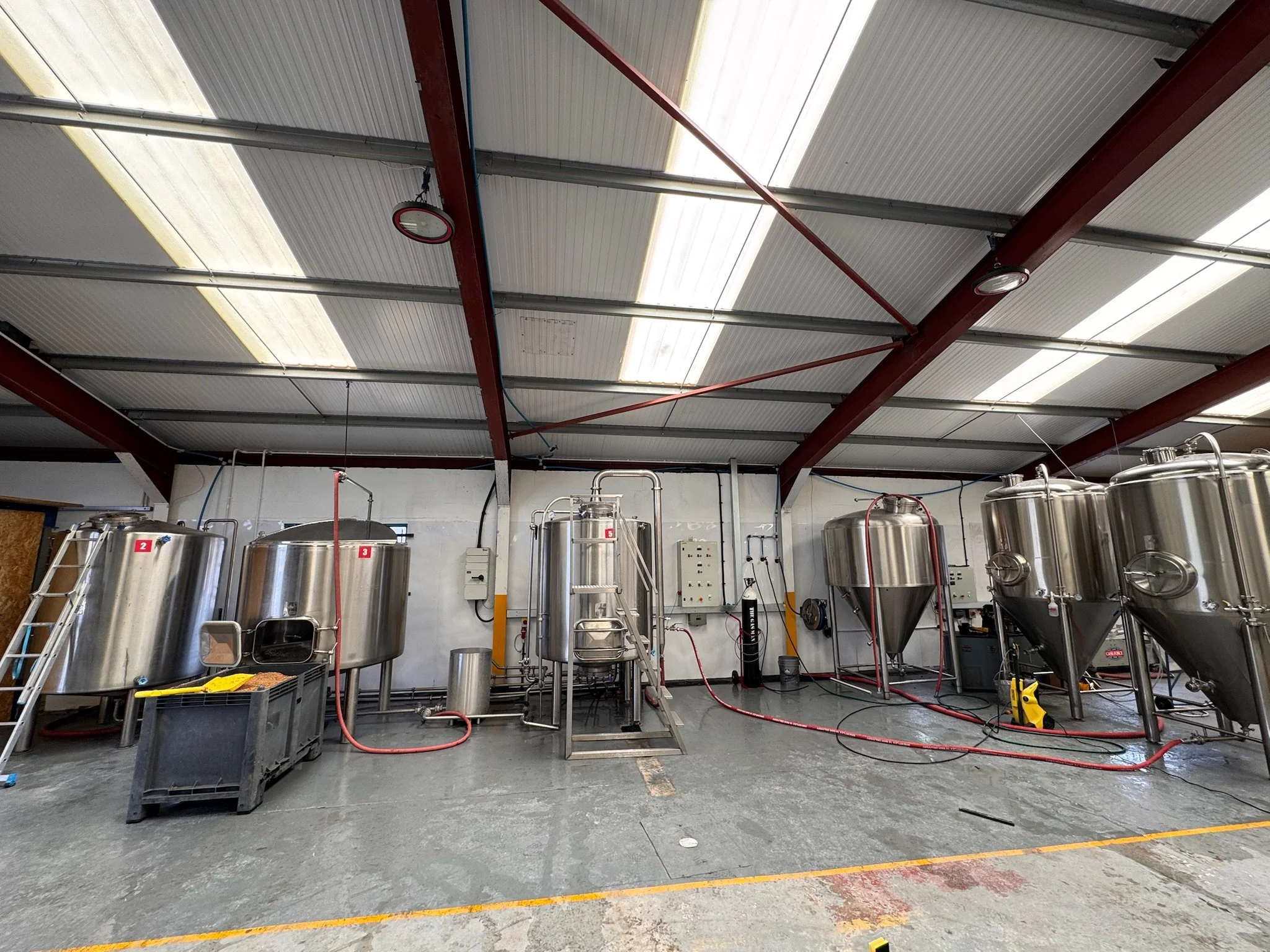 Industrial brewery room with stainless steel fermentation tanks connected by hoses and pipes, and various equipment against a concrete wall and metal ceiling.