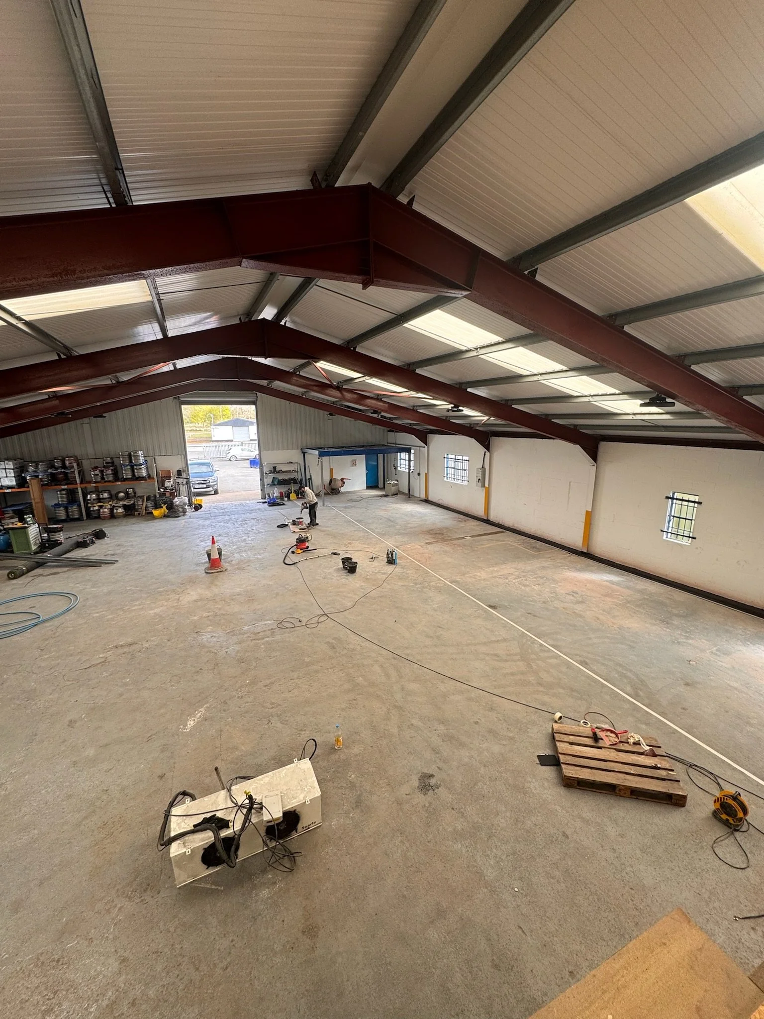 Inside view of a large industrial building with a high ceiling, visible steel beams, and a concrete floor, where construction or renovation work is taking place. There are tools, a pallet, and a worker in the background.