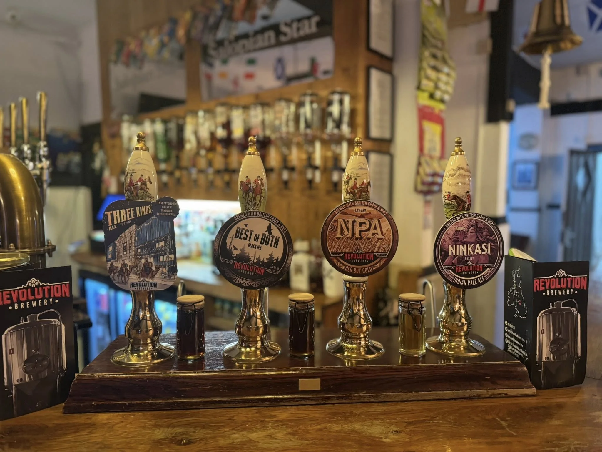 Five beer taps from Revolution Brewery with colorful labels and small glass bottles on a wooden bar counter, with a bar decor background.