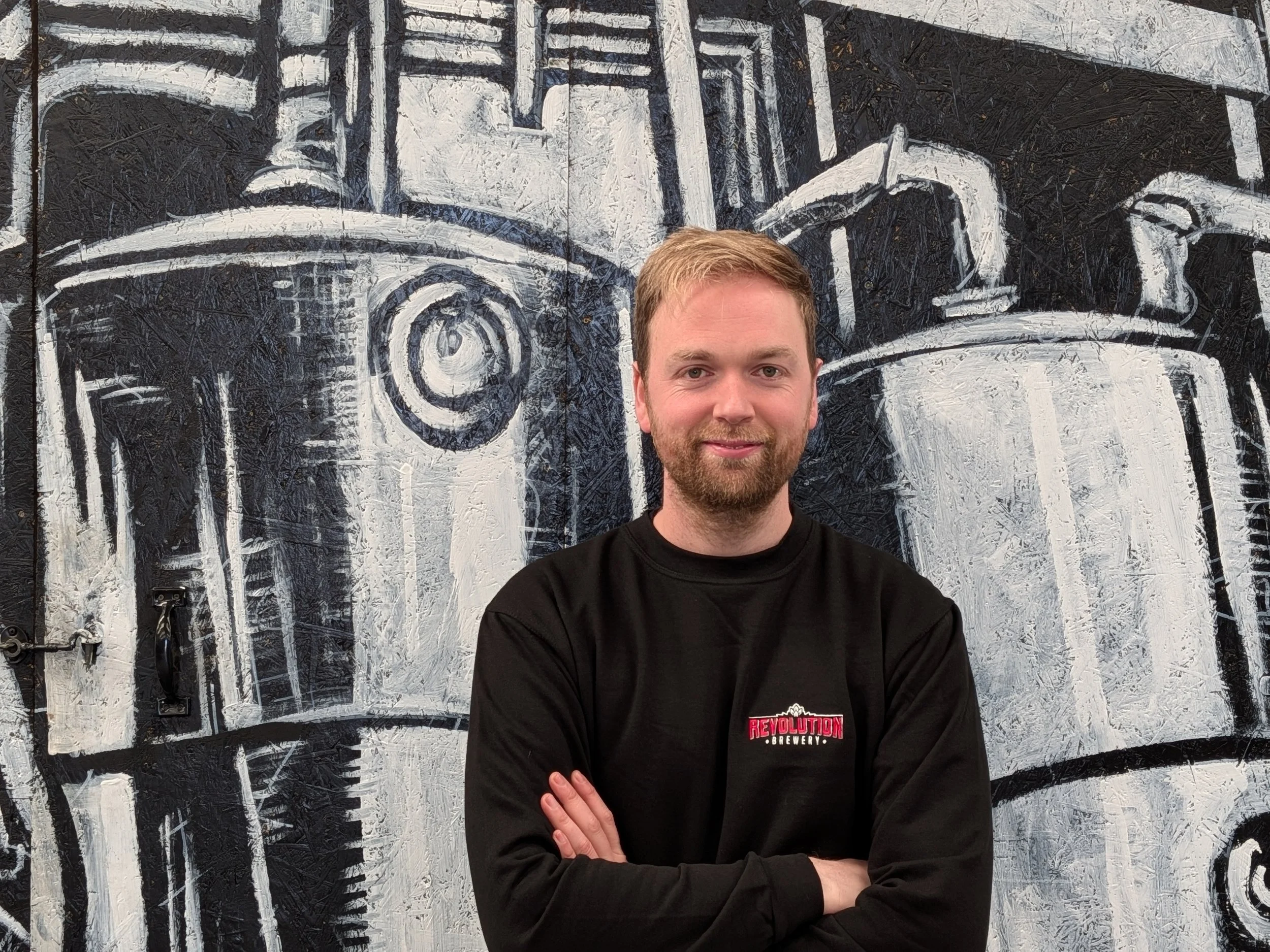A man with a beard and blonde hair, wearing a black sweatshirt with a red and white logo, standing with arms crossed in front of a black and white mural of vintage streetcars.