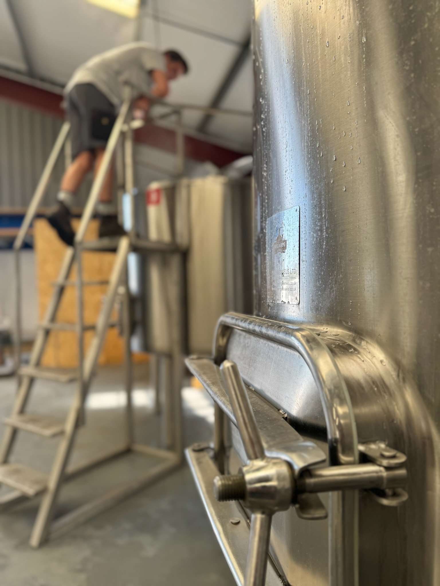 Close-up of a metallic container with a valve and lever, with a person on a ladder in the background working on another container in an industrial setting.