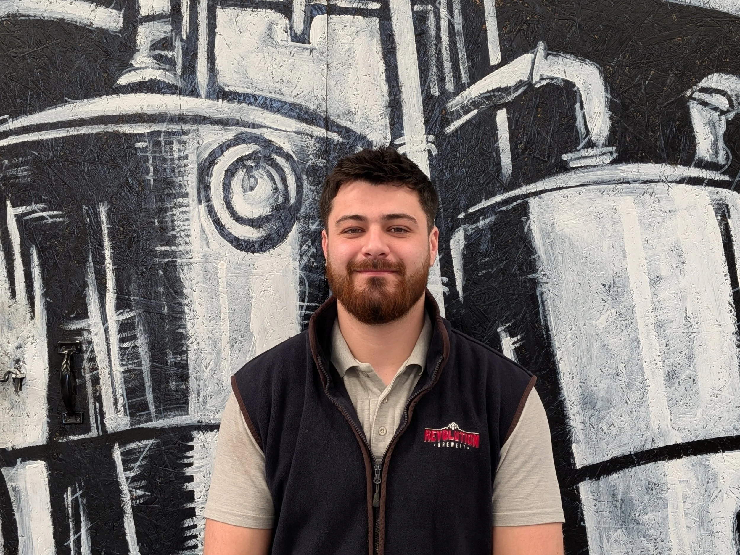 A young man with a beard and short dark hair standing in front of a black and white industrial-style mural of a large machine or engine. He is wearing a beige polo shirt and a black vest with red embroidery.