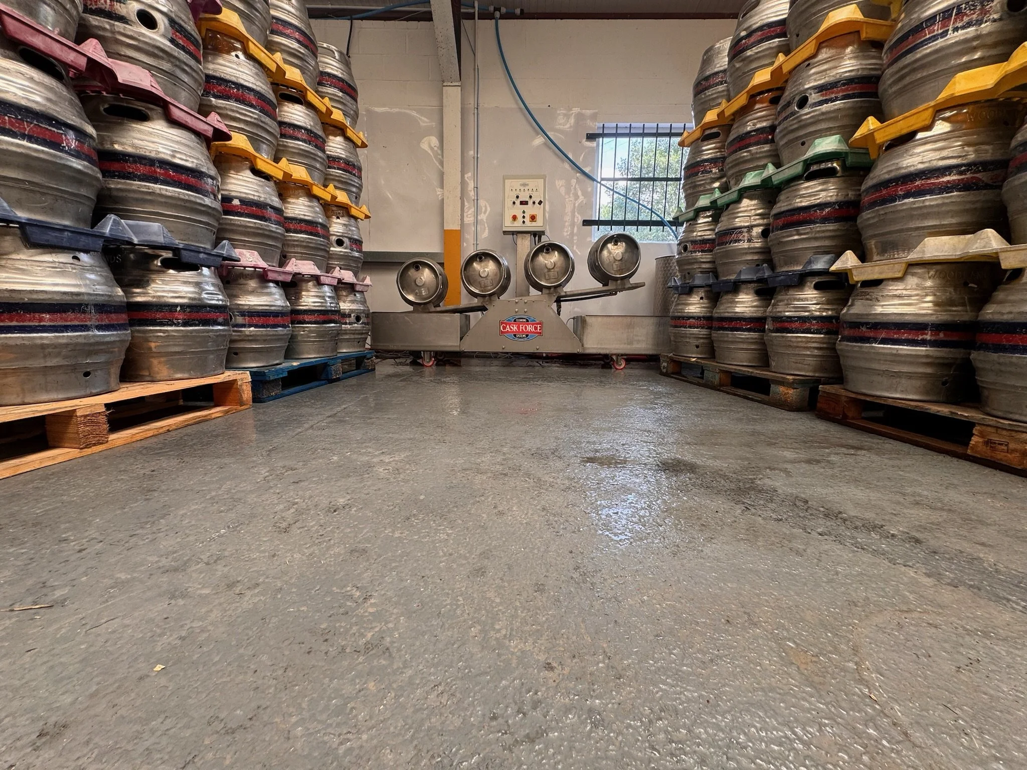 Storage room with stacks of empty beer kegs on pallets on both sides and a machine in the center used for filling or cleaning kegs, with a window in the background letting in natural light.