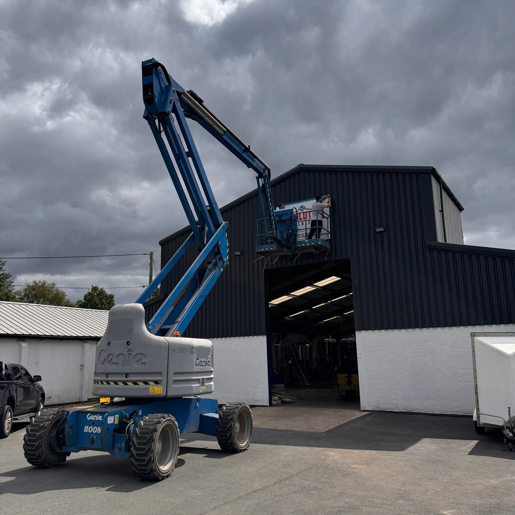 Two workers on a blue boom lift painting or installing a sign on a dark gray building's upper section.