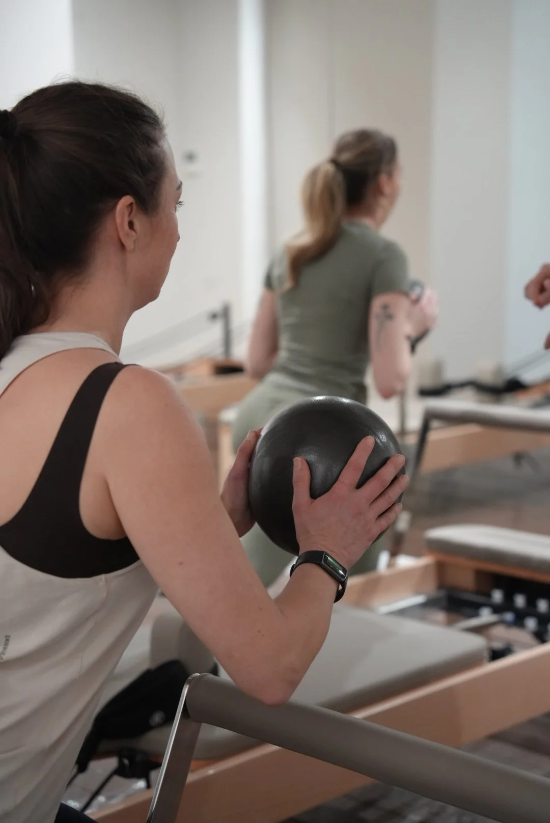A woman in workout clothes holding a black medicine ball during a fitness class, with other participants in the background.