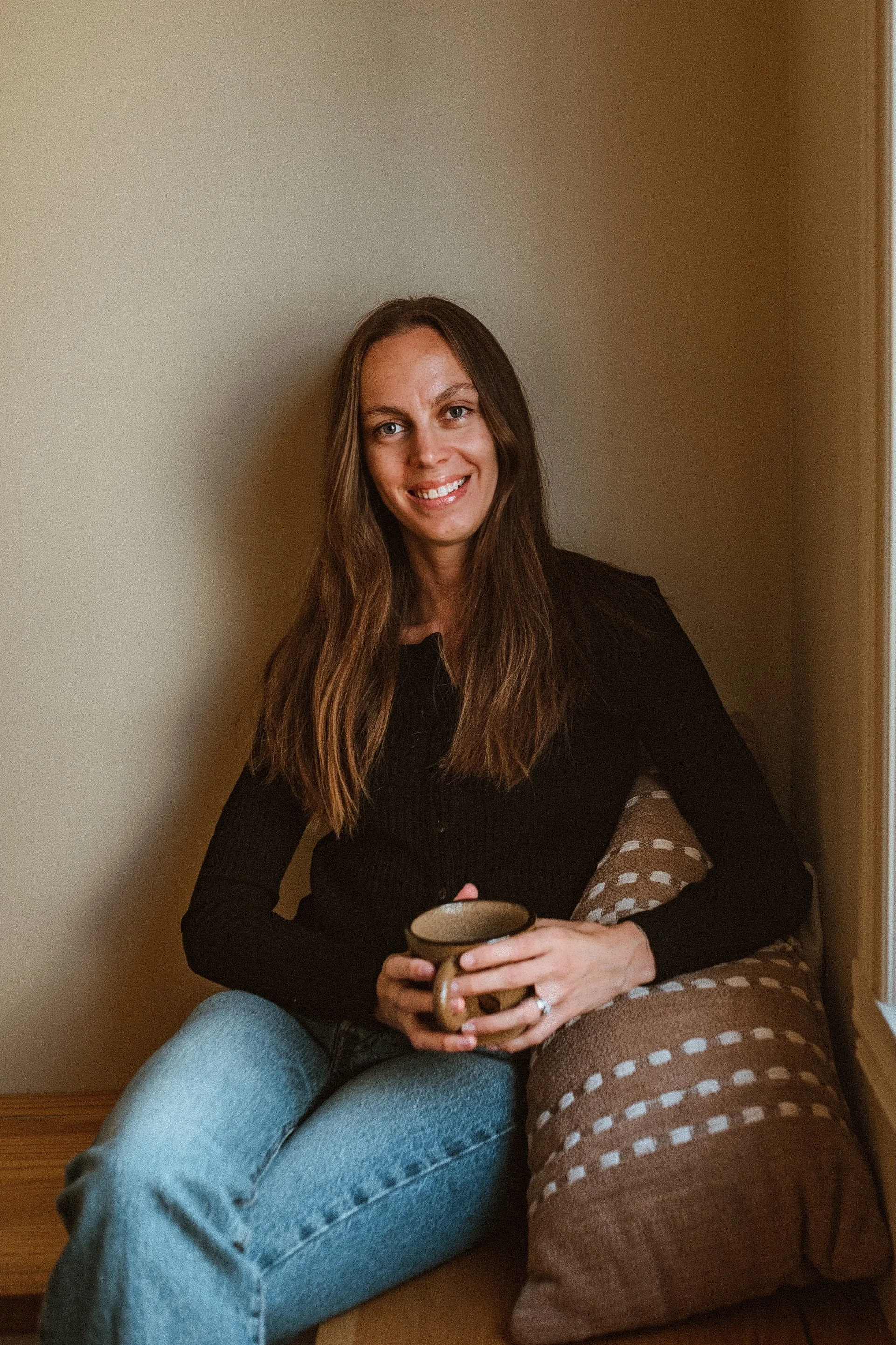 Femme souriante assise près d'une fenetre, tenant une tasse.