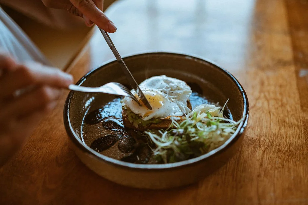 Une personne casse un œuf dans une assiette en céramique contenant une tranche de pain, de la laitue et de la sauce.