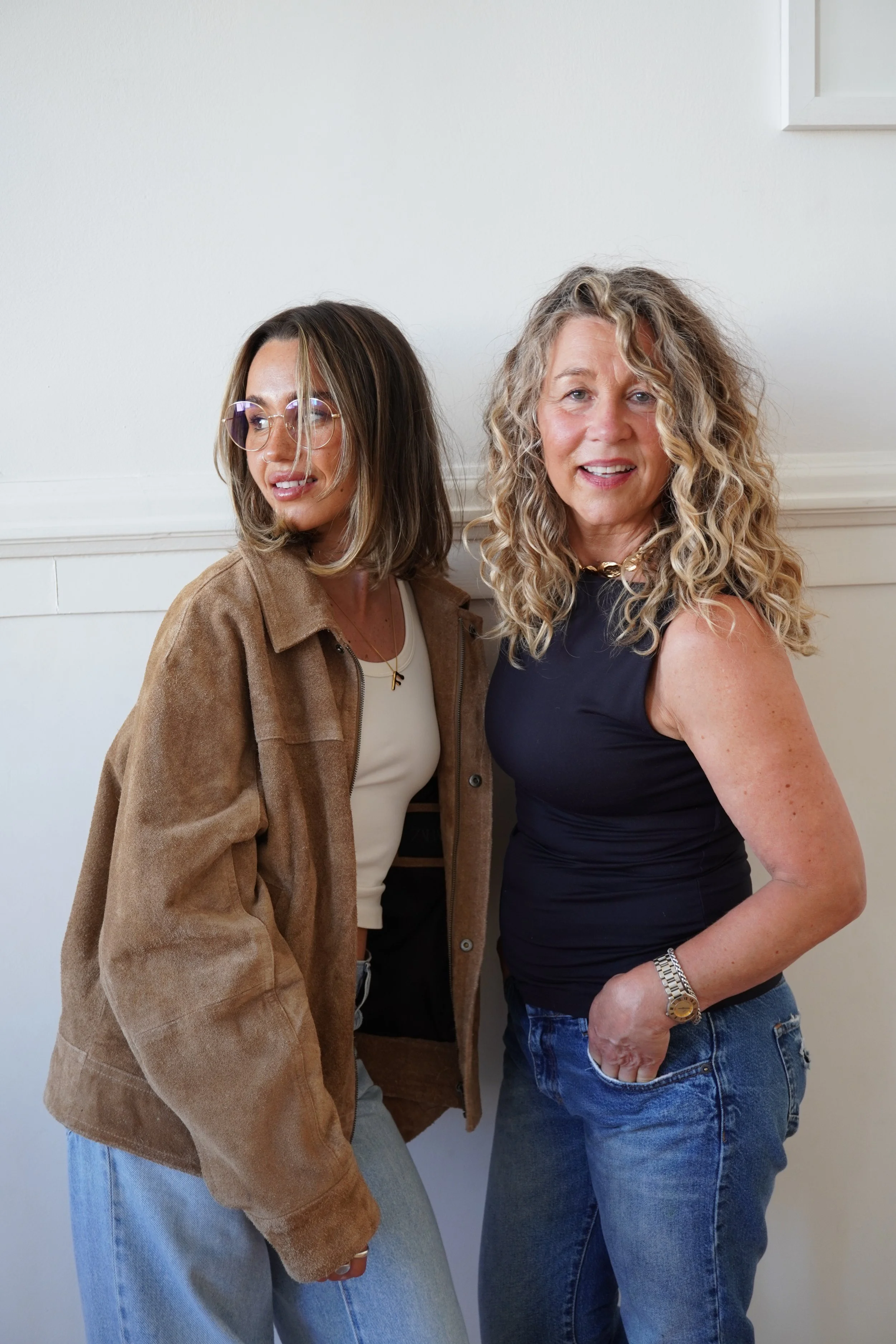 Two women standing next to each other indoors against a white wall, one wearing glasses and a brown jacket, the other with curly blonde hair and a black sleeveless top, both smiling at the camera.