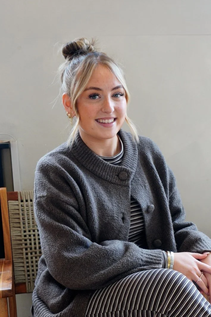 A young woman with blonde hair styled in loose waves, smiling and standing indoors in front of wooden shelves displaying skincare products. She is wearing a cream-colored knit sweater with black horizontal stripes and dark pants.