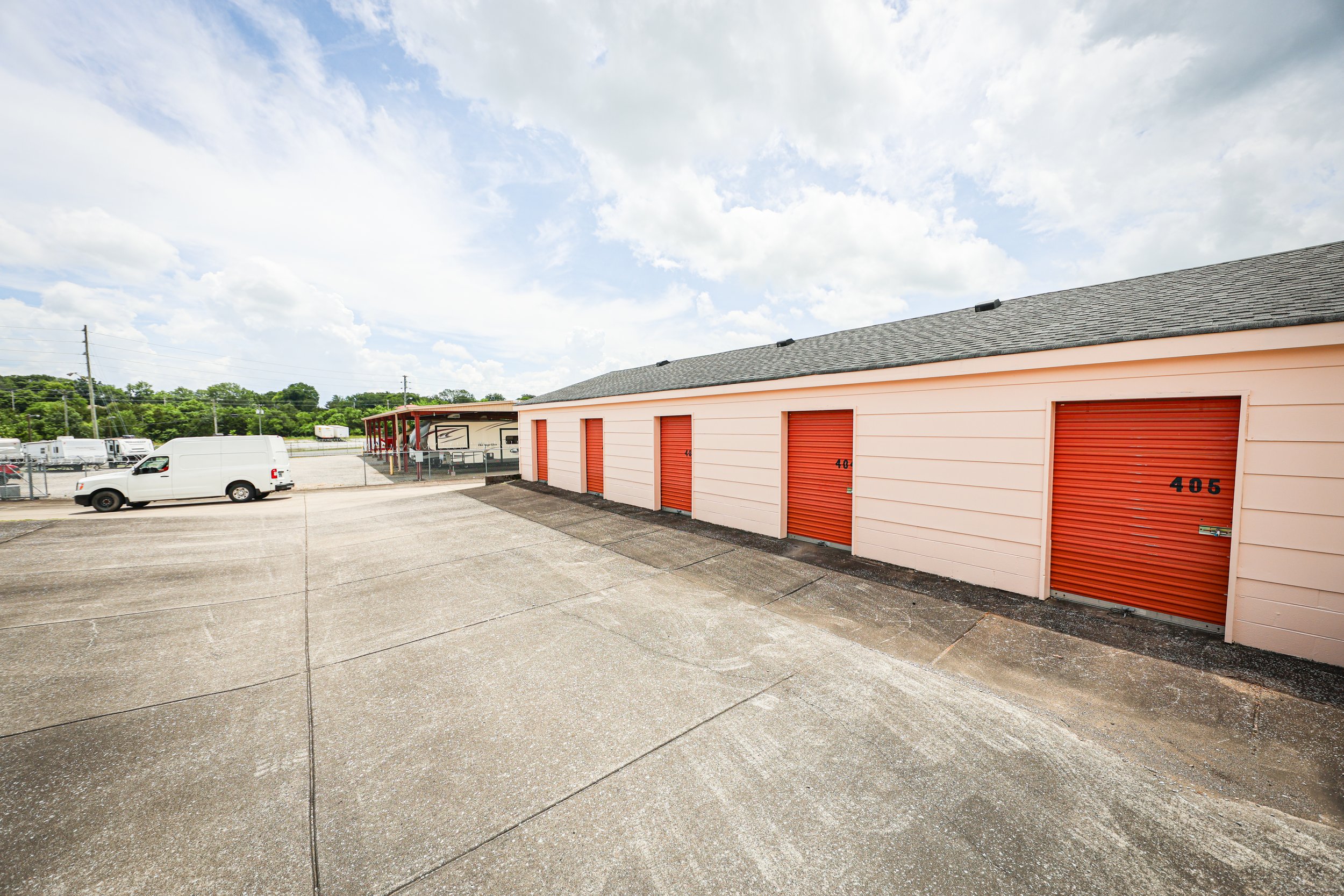 David Jent Mini-Storage facility with several orange roll-up doors, numbered 404, 405, and others, in a paved lot with a white van and a covered area with recreational vehicles in the background.