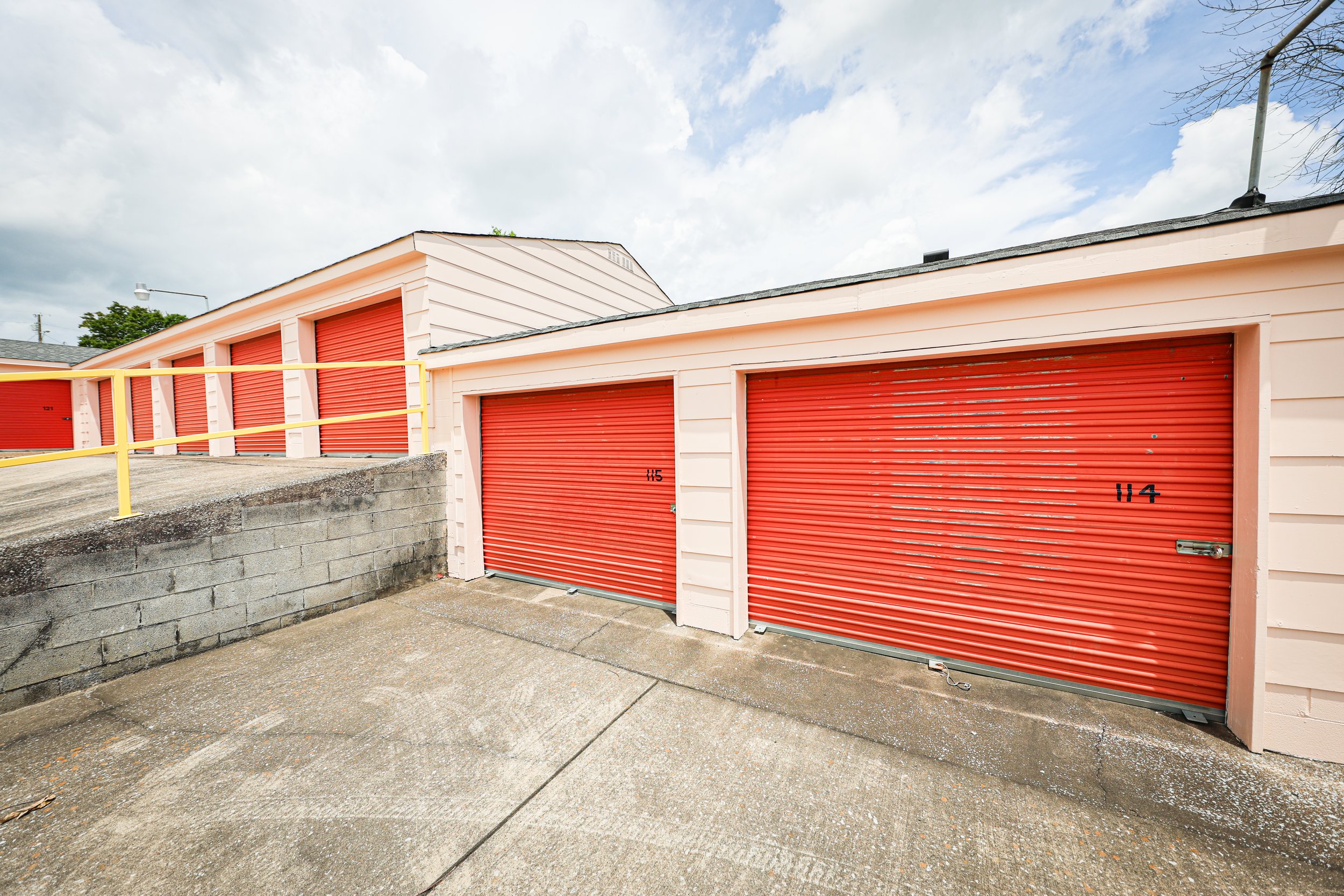 David Jent Mini-Storage. Row of storage units with orange metal roll-up doors, numbered 114 and 115, in an outdoor storage facility.