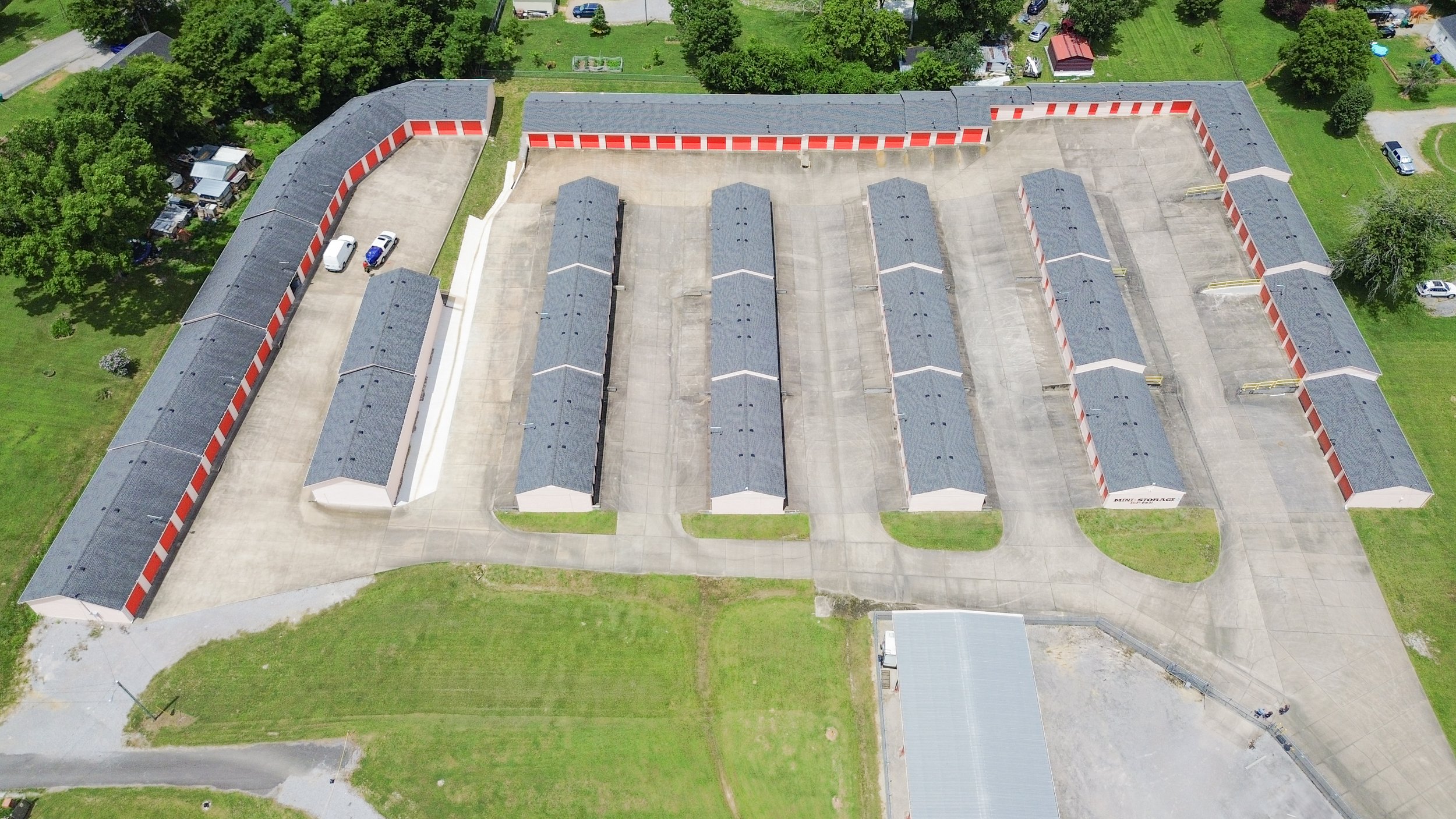 An aerial view of a David Jent Mini-Storage facility with multiple rows of storage units, some with red doors, surrounded by green lawns and trees.