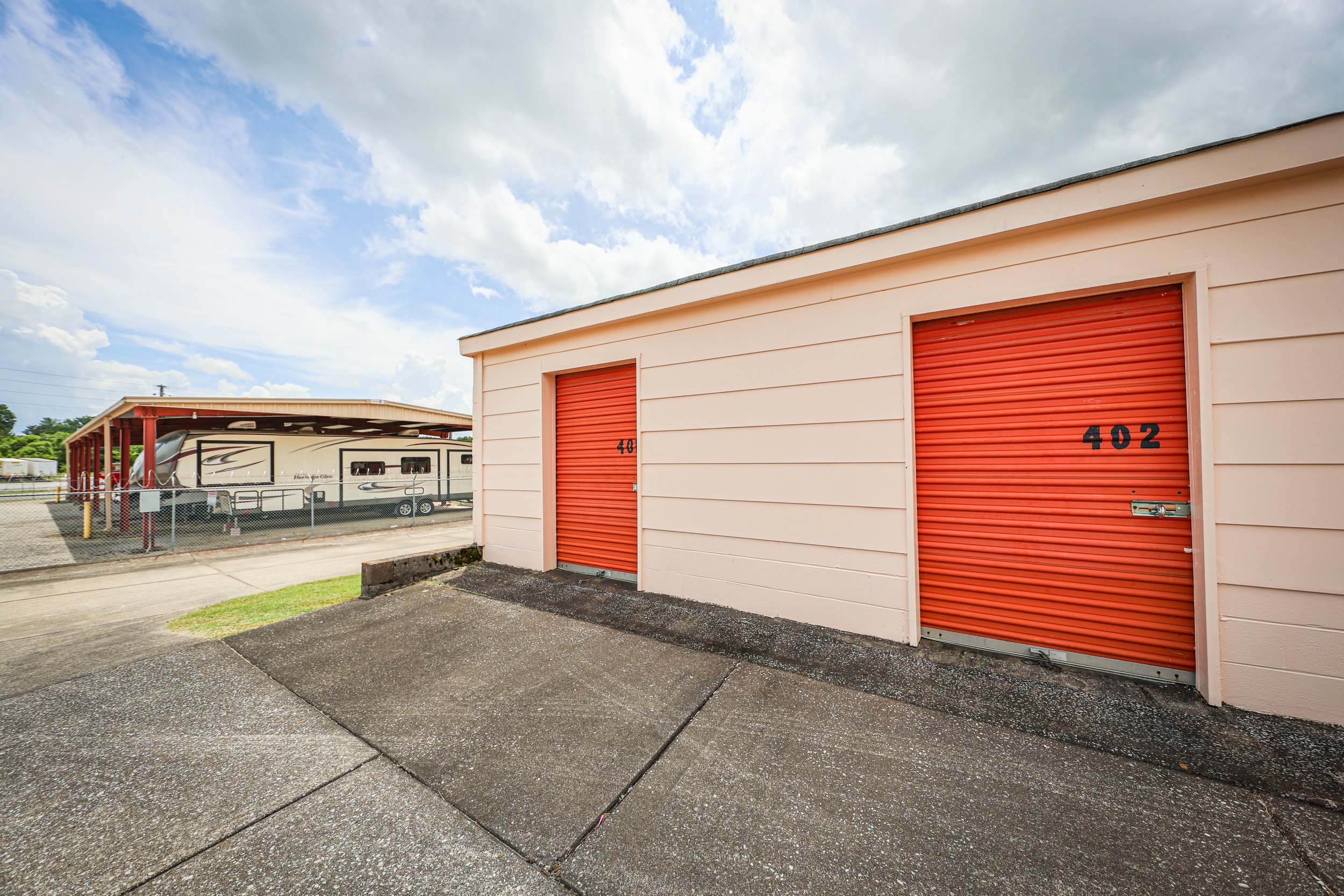 Exterior view of a David Jent Mini-Storage facility with two orange roll-up doors numbered 402 and 403, on a concrete surface, with a recreational vehicle parked under a red-roofed carport in the background, and a partly cloudy sky overhead.