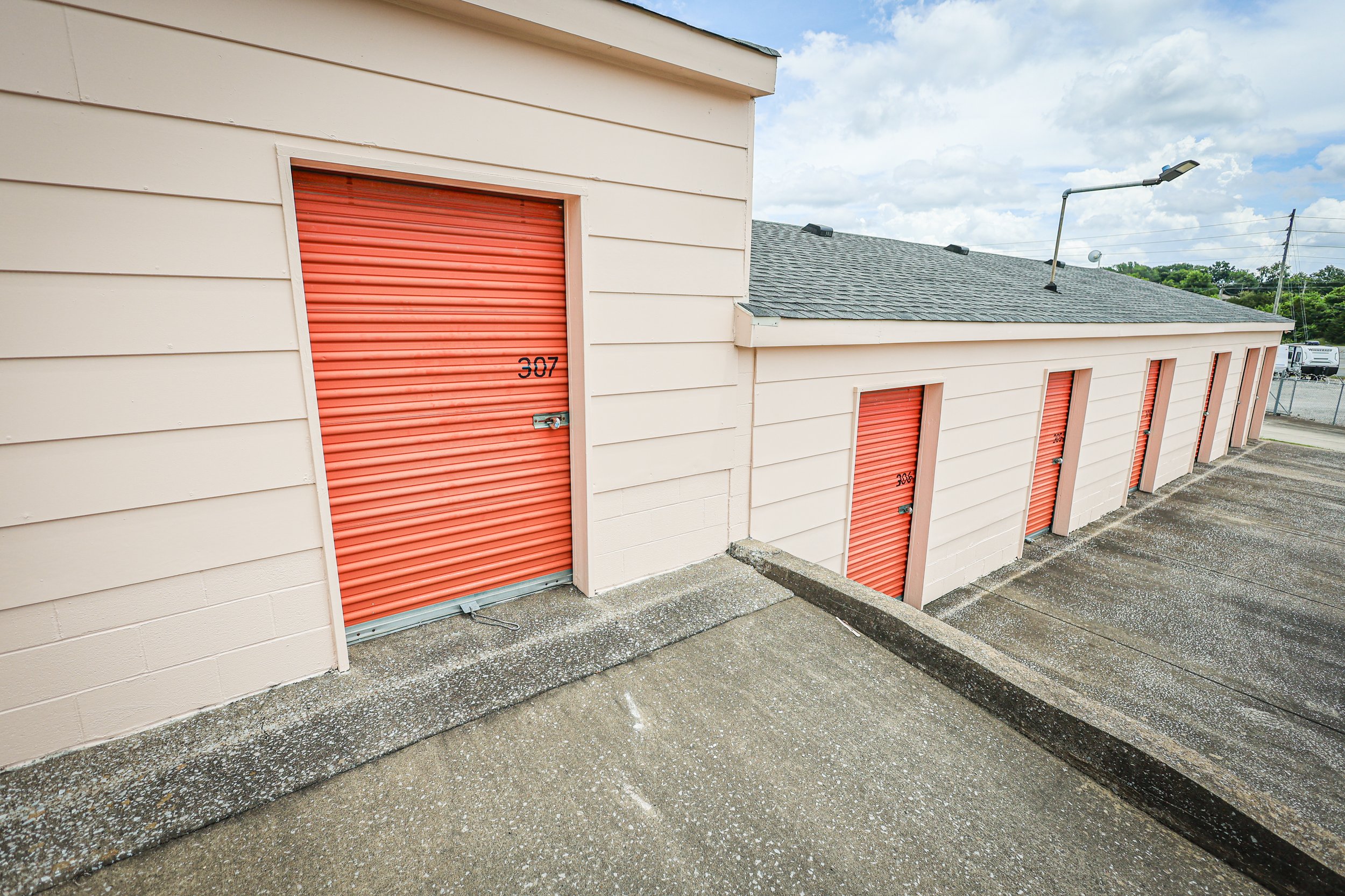 A row of storage units at David Jent Mini-Storage with orange roll-up doors on a concrete rooftop in daytime, numbered 307, 305, 306, 304, 303, and 302, with a cloudy sky and a street with cars and streetlights in the background.