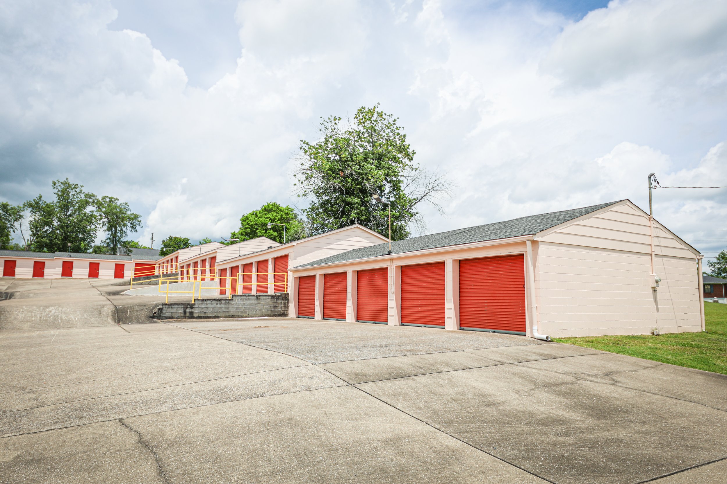 A row of storage units with orange roll-up doors, some with small buildings and a yellow guard rail in front, under a partly cloudy sky.