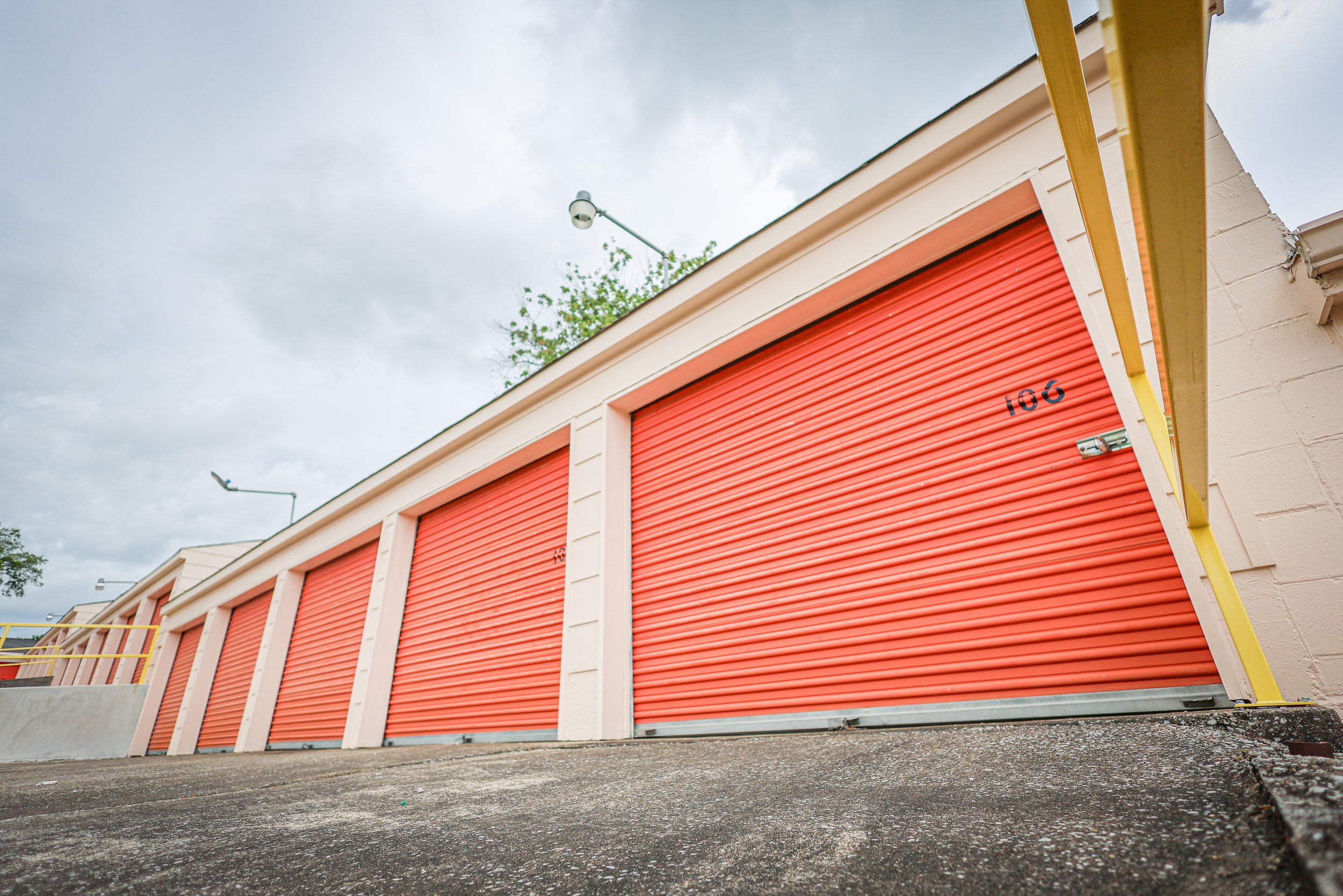 A row of orange storage unit roll-up doors at David Jent Mini-Storage with yellow piping along the top, viewed from a low angle on a cloudy day.