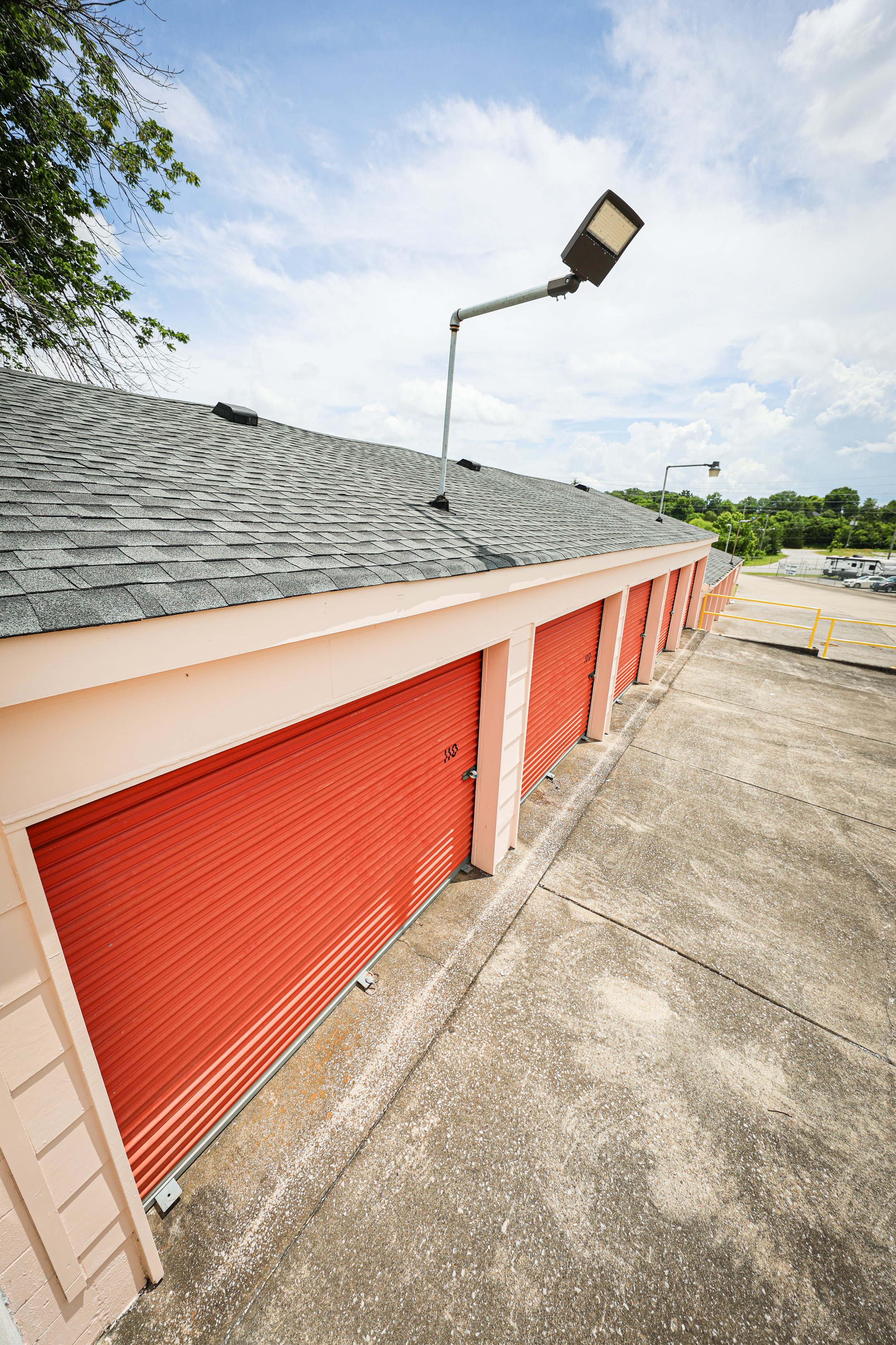 David Jent Mini-Storage. Rows of orange storage unit garage doors on a rooftop, with a gray shingled roof and a large outdoor light fixture against a partly cloudy sky.