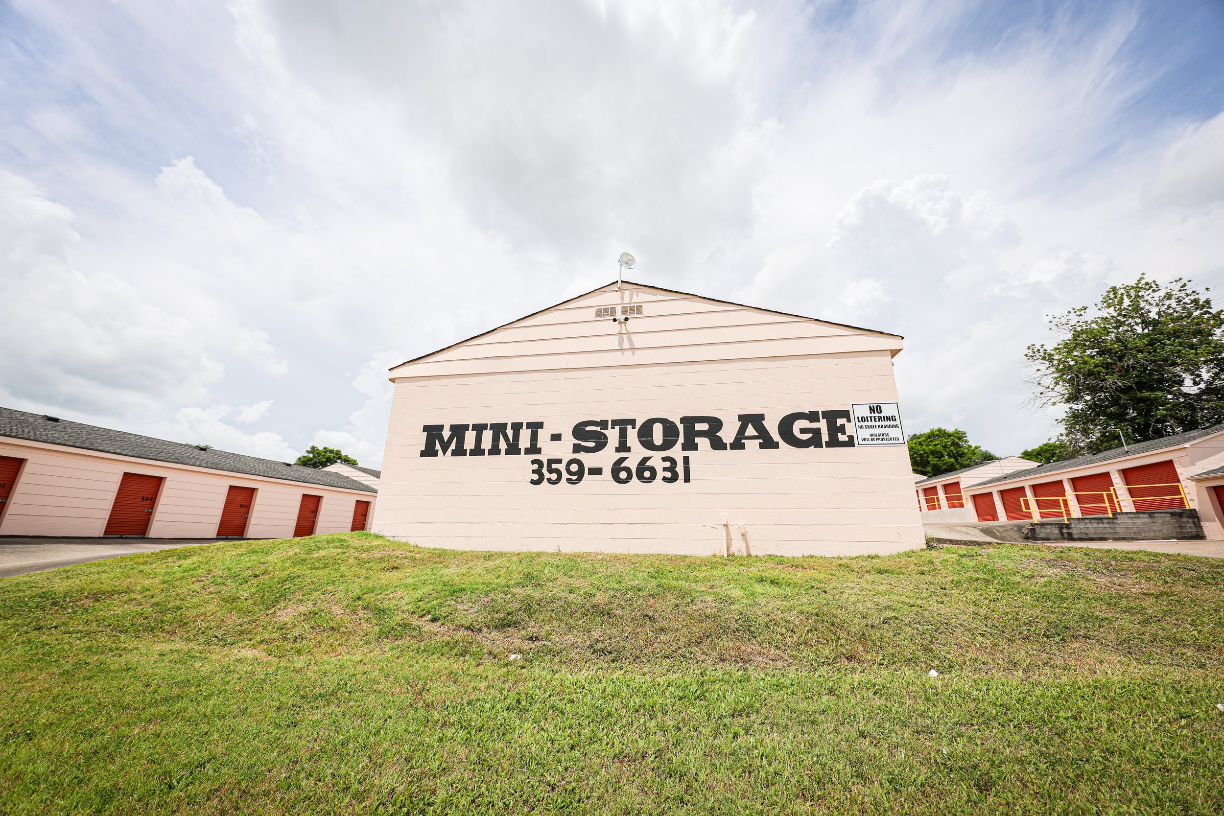 David Jent Mini-Storage. Smaller storage units with orange doors surrounding the building, and a grassy area in front. The sky is partly cloudy.