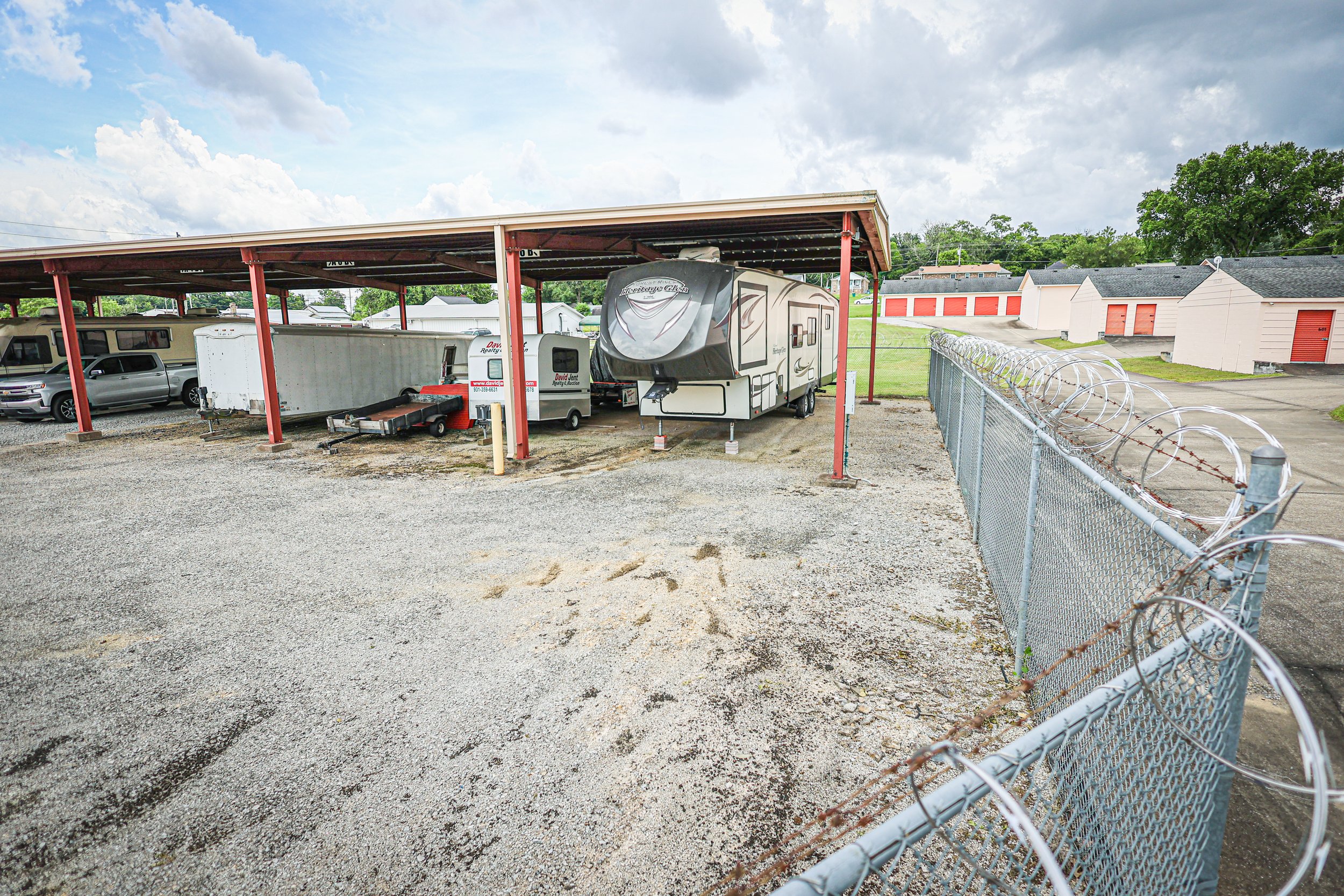 A fenced outdoor storage lot at David Jent Mini-Storage with RVs and trailers parked under a metal shelter, barbed wire on the fence, and a row of garages in the background.