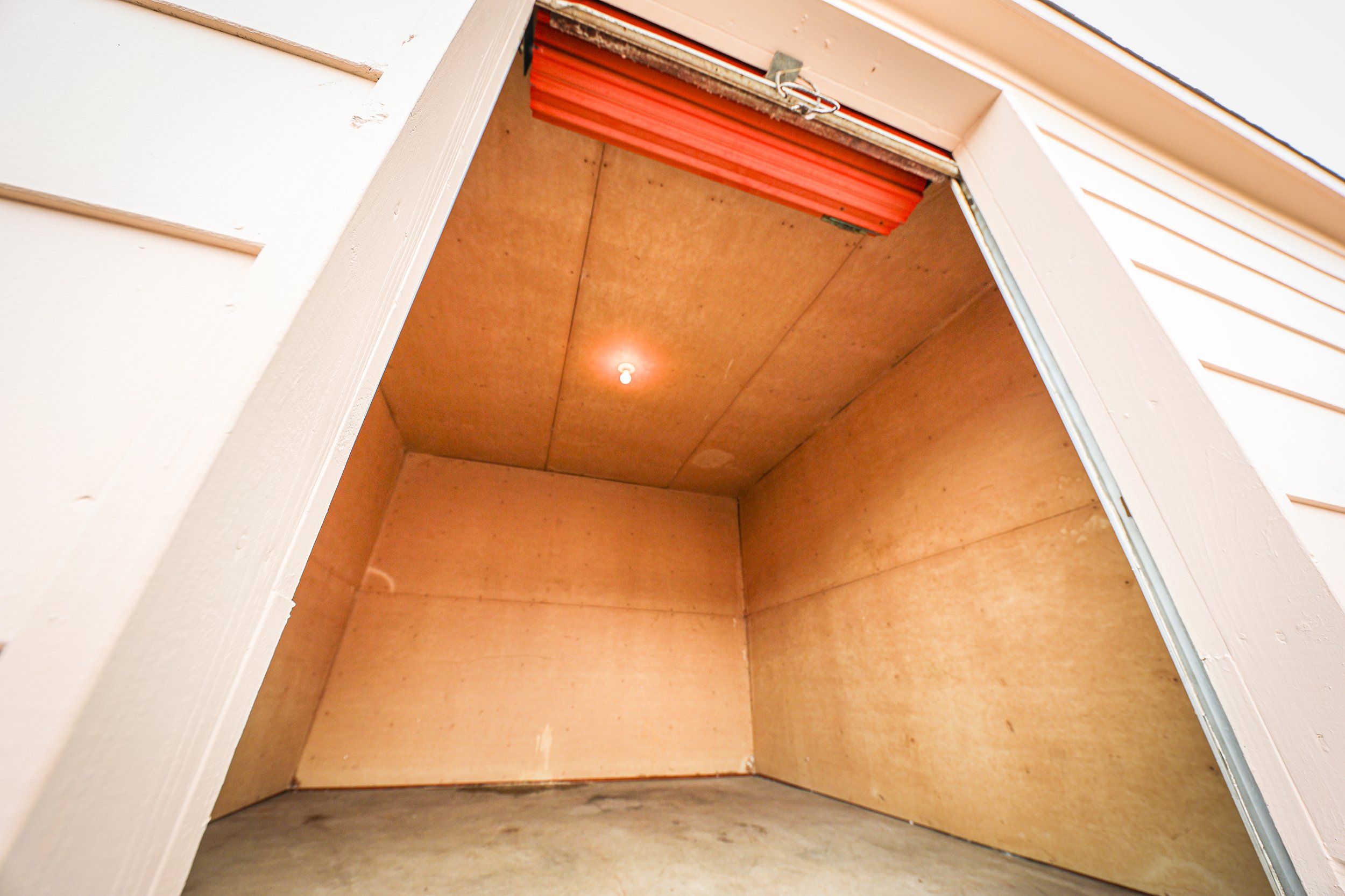 Empty storage unit at David Jent Mini-Storage with an open doorway, interior walls and ceiling made of plywood, concrete floor, and a closed orange roll-up door at the back.