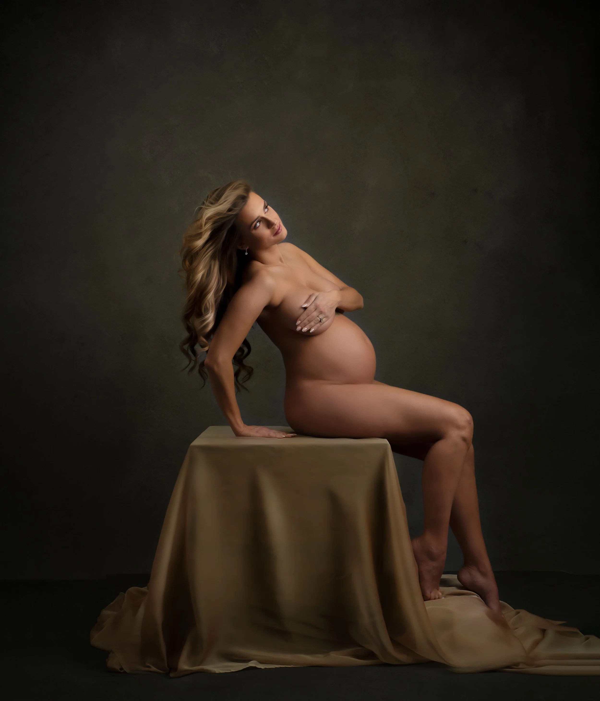 A nude woman with long wavy hair sitting on a draped table against a dark background, with one hand covering her chest and the other supporting her on the table, gazing upward.