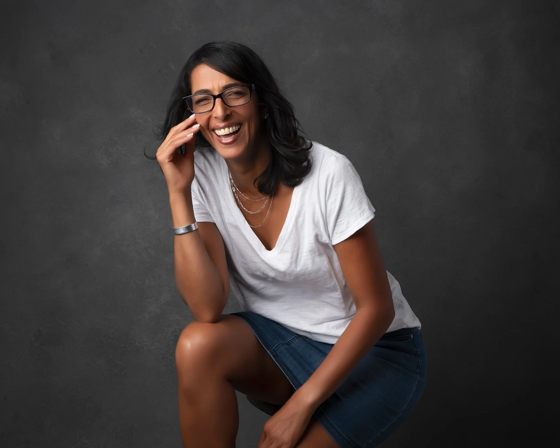 Woman with black hair and glasses, smiling and laughing, seated against a dark grey background, wearing a white t-shirt and denim skirt.