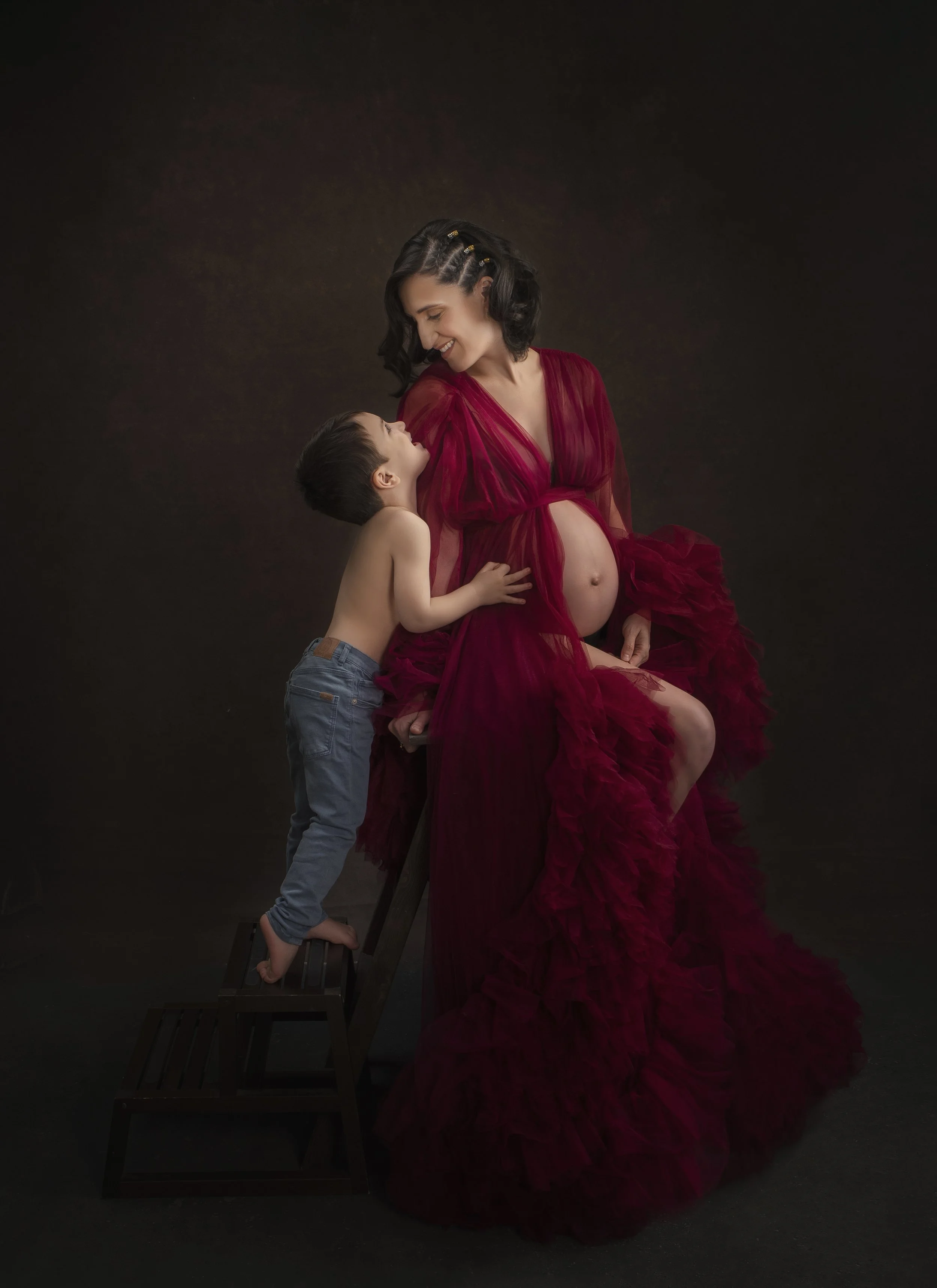 A pregnant woman in a flowing red dress sitting on a stool, smiling at a young boy who is shirtless and wearing blue jeans standing on a small wooden step stool, reaching up to her, against a dark background.