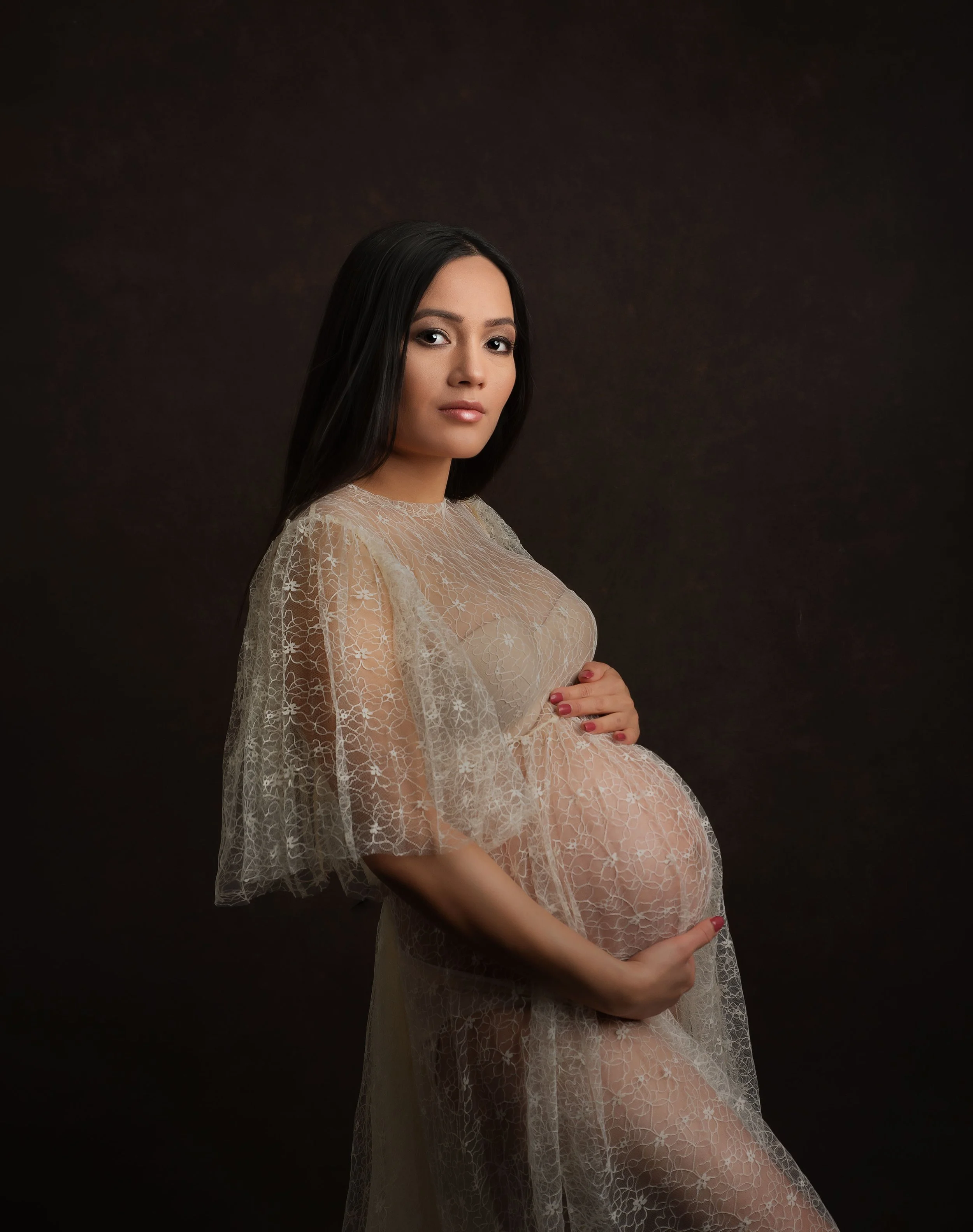 A pregnant woman with long dark hair wearing a sheer, floral lace dress holding her belly against a dark background.