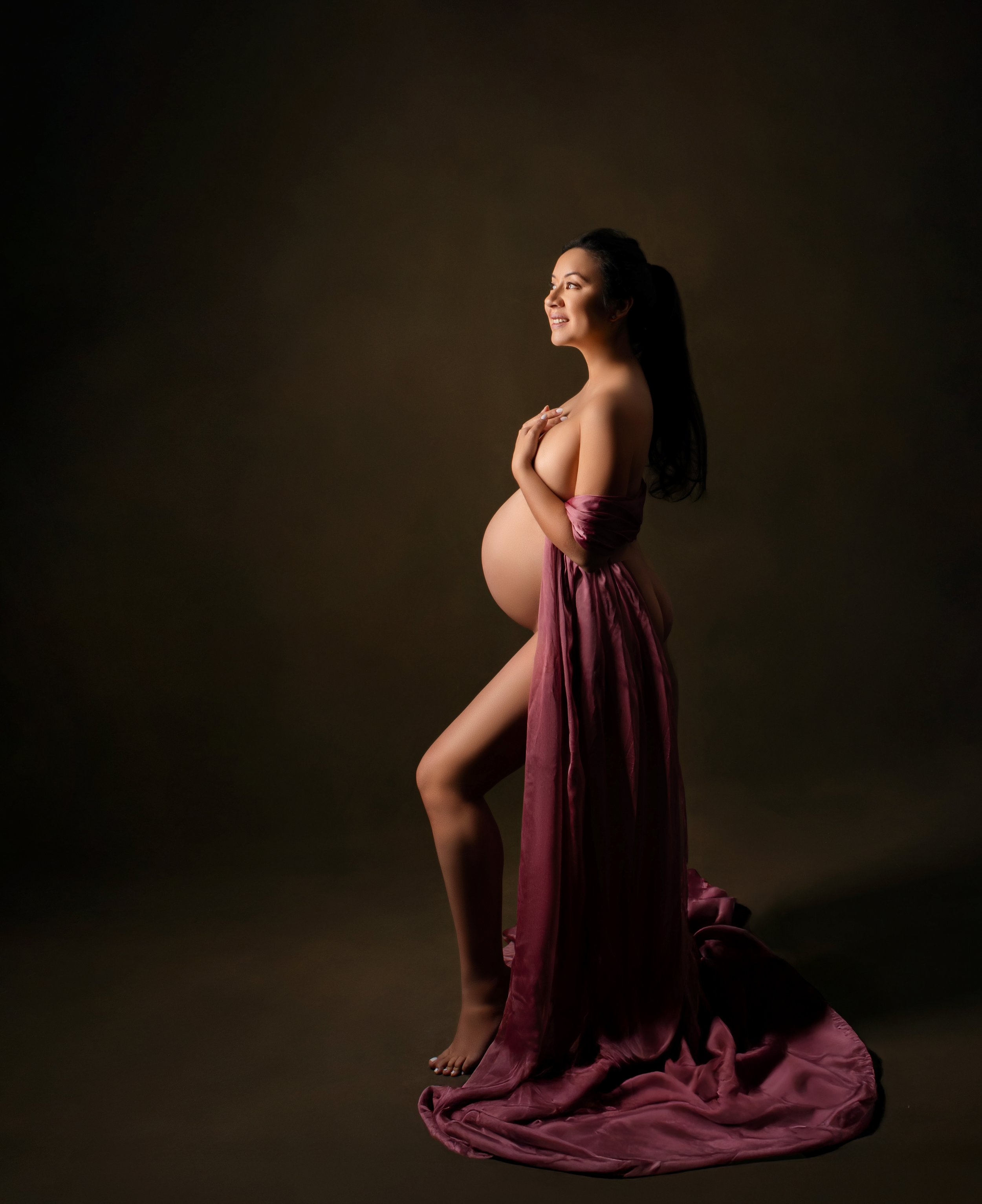 Pregnant woman in a pink gown standing on a dark background, holding her belly and smiling.