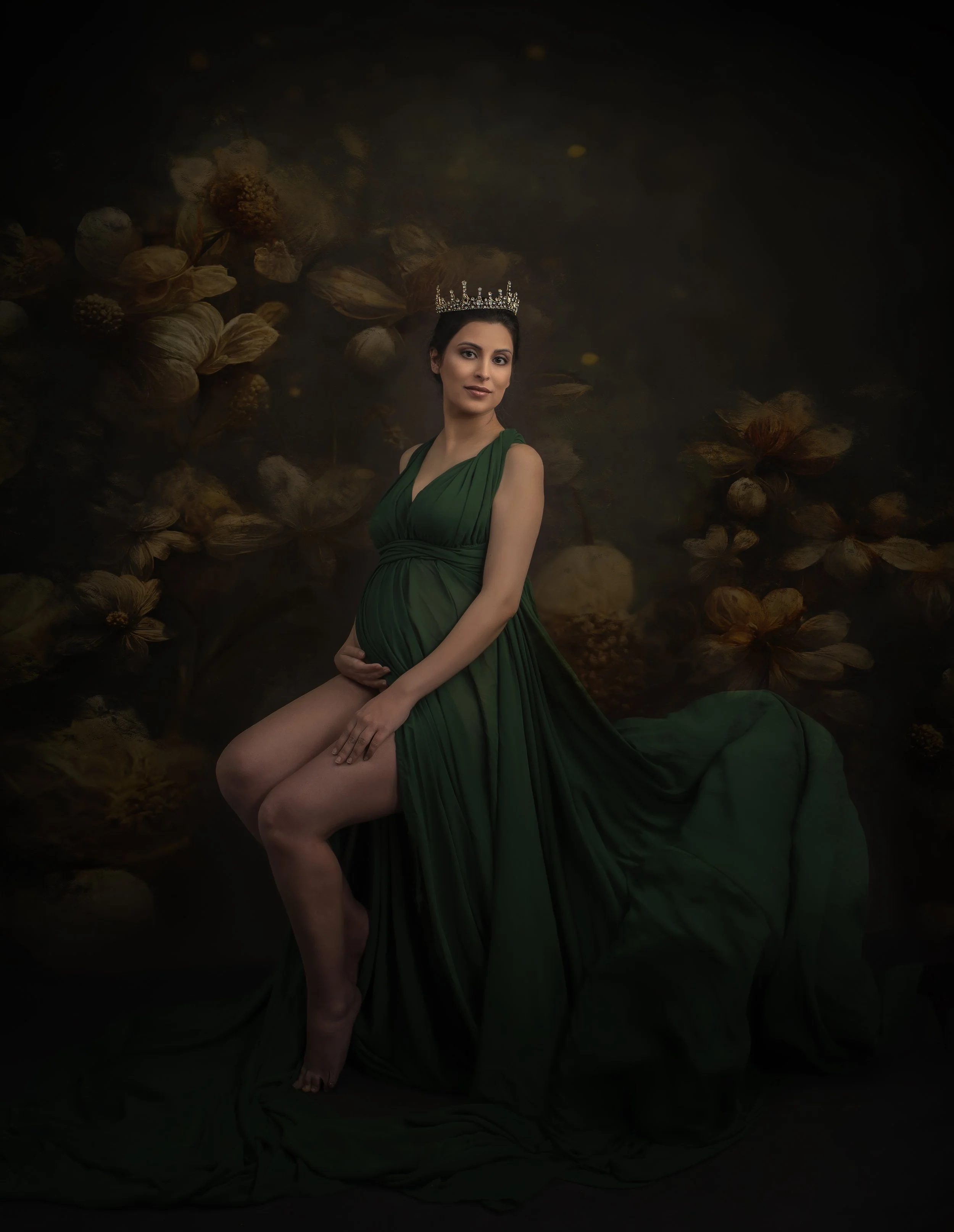 A pregnant woman wearing a flowing green dress and a tiara, sitting with one knee raised and the other leg extended on a dark background with dried flowers.