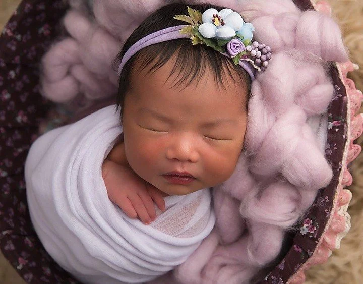 Photoshoot by Ally, A sleeping baby girl with a floral headband, swaddled in white cloth, lying on pink cotton, inside a round basket lined with pink fabric.