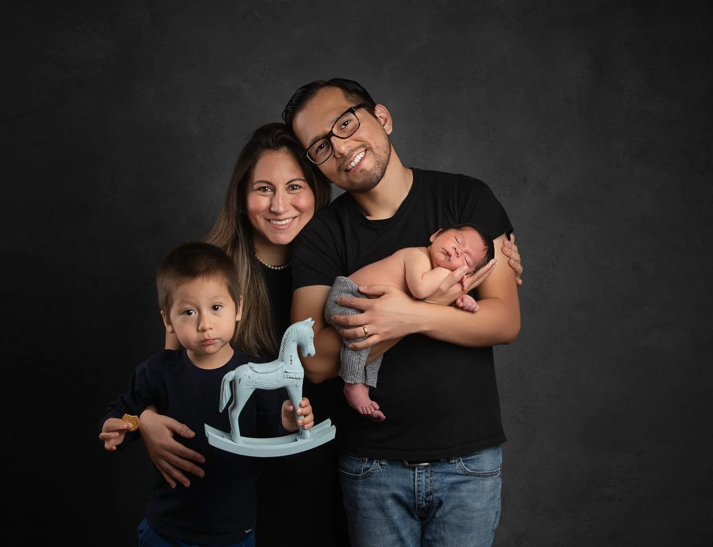 Photoshoot by Ally, Family portrait of four with a mother, father, and two children, one holding a baby. The father is holding the baby, who is sleeping, while the mother and older son stand nearby, with the son holding a toy horse.