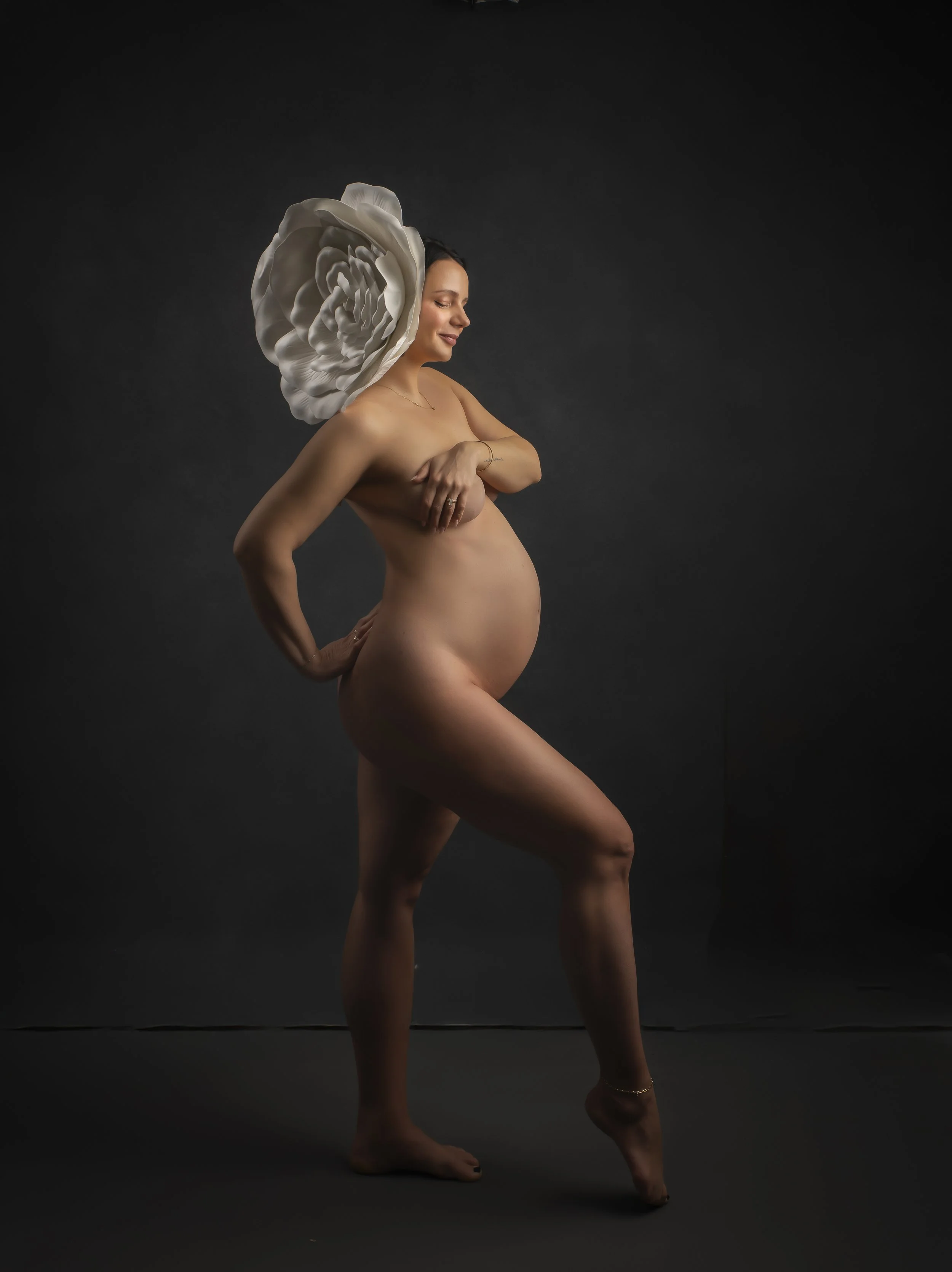 A nude pregnant woman with a large white flower on her head, standing against a dark background, smiling with her eyes closed.