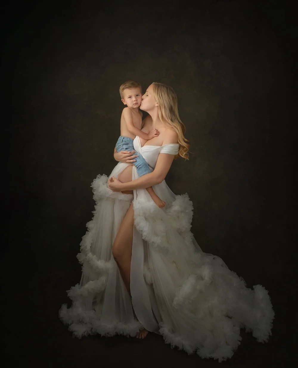 A woman in a white dress holding a young boy, who is shirtless and wearing jeans, against a dark background. The woman is kissing the boy on the cheek.