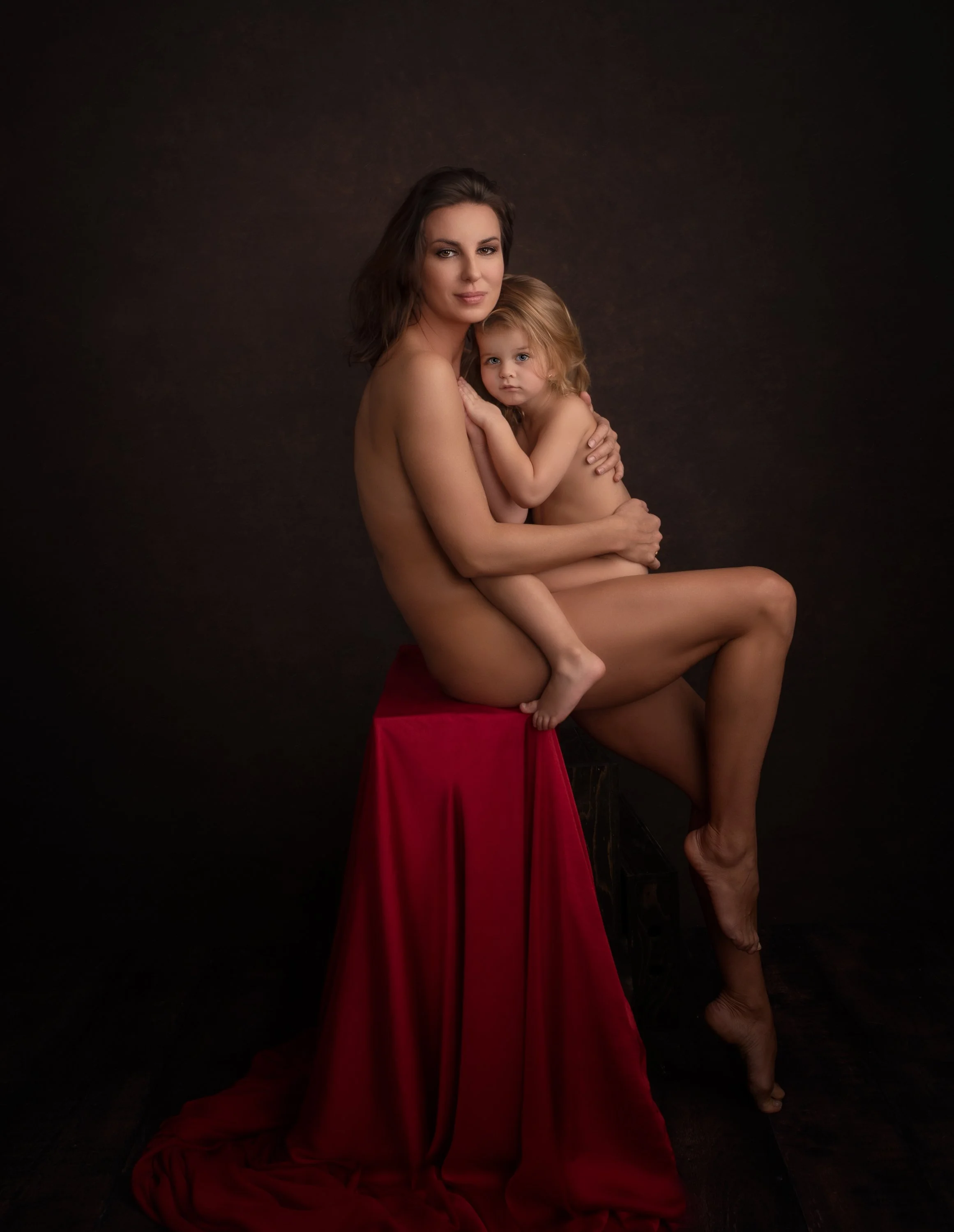 A woman and a young girl, both nude, sitting on a red cloth-covered stool against a dark background, embracing each other.