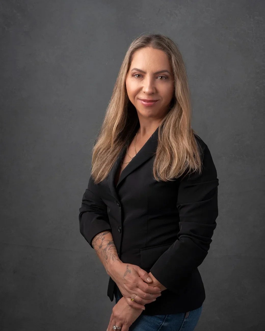 Photoshoot by AllyA woman with long blonde hair, wearing a black blazer and blue jeans, standing against a gray background, smiling slightly and looking at the camera.
