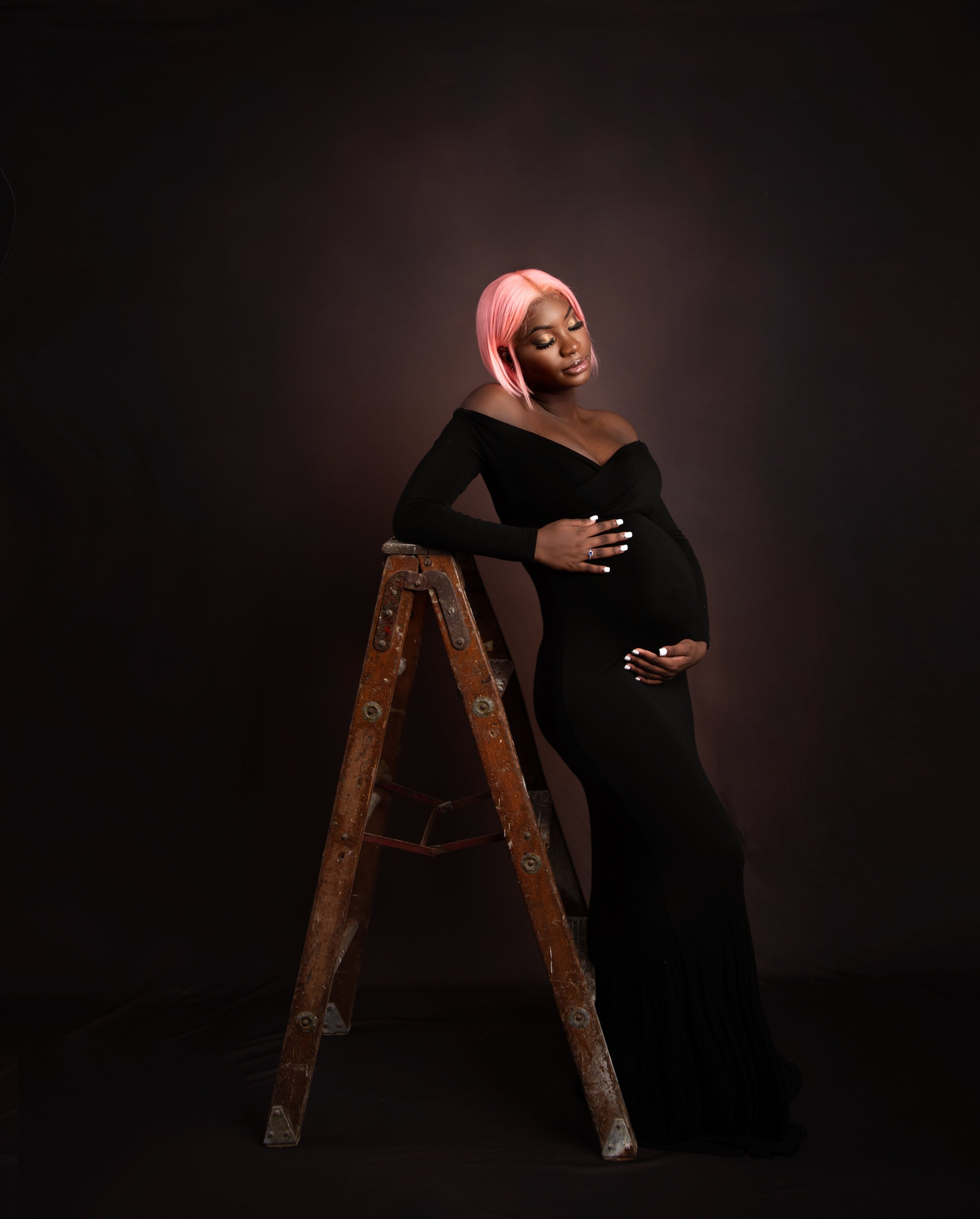 Pregnant woman with pink hair in a black dress leaning on a ladder against a dark background.