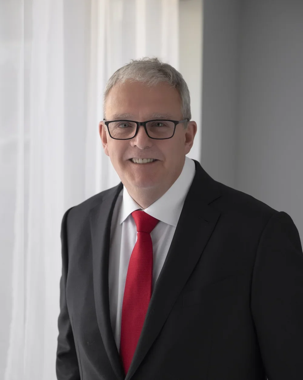 Photoshoot by Ally, A middle-aged man wearing glasses, a black suit, white shirt, and red tie, smiling and standing indoors near sheer curtains with natural light.