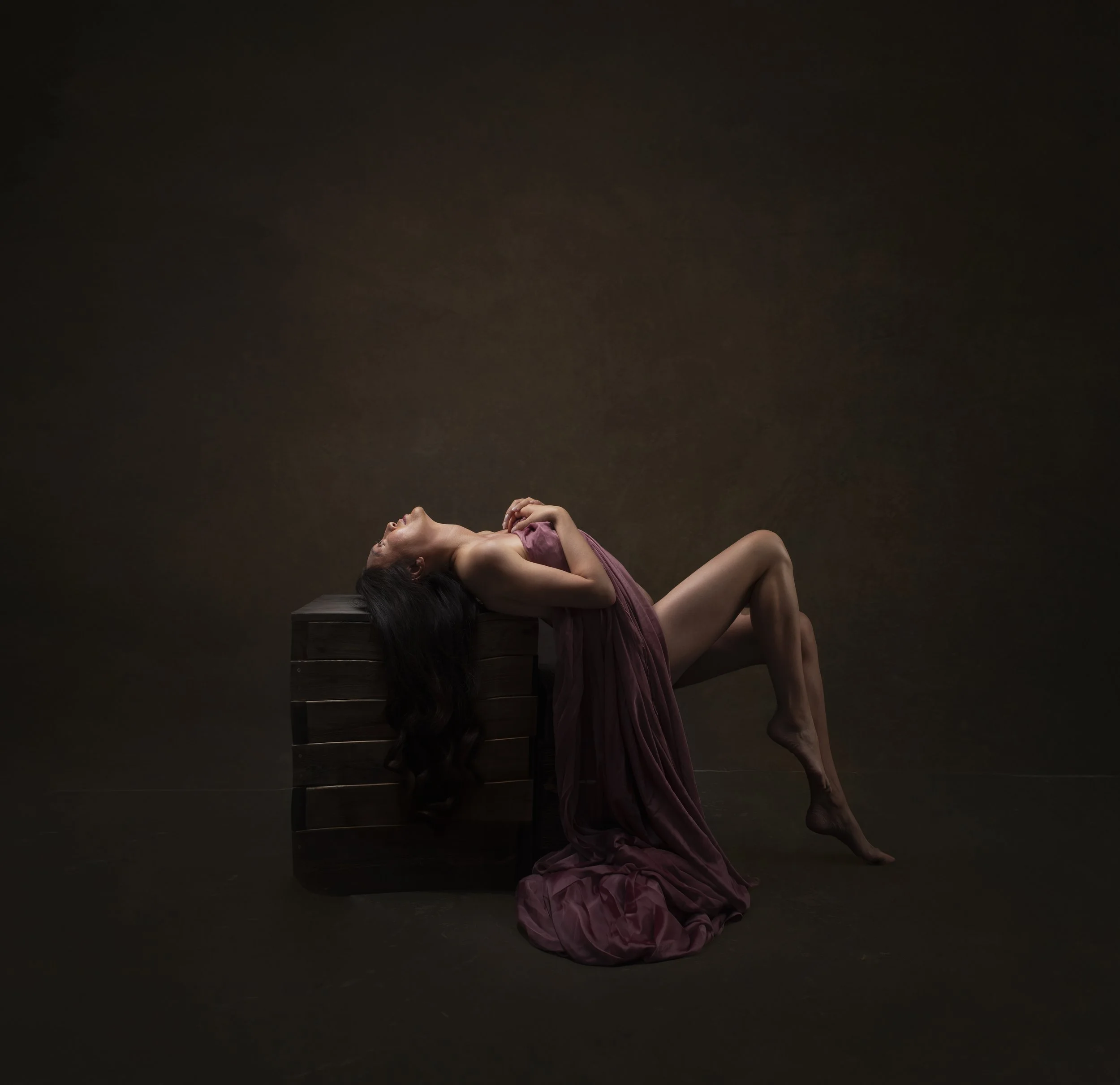A woman with long dark hair reclining on a wooden box, draped in a purple fabric, with a dark background.
