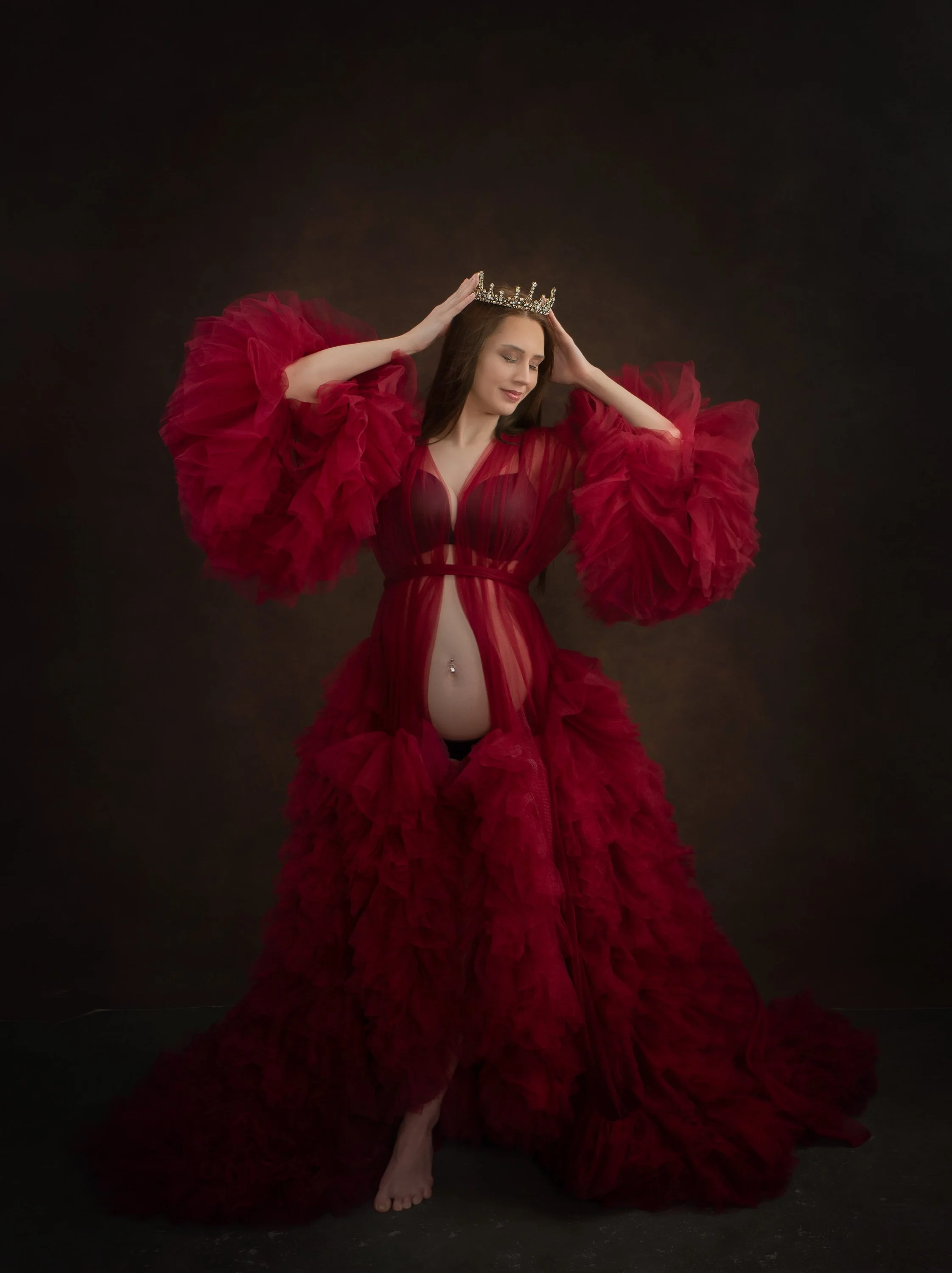 A woman in a red, ruffled dress with puffed sleeves holds a crown on her head against a dark background.