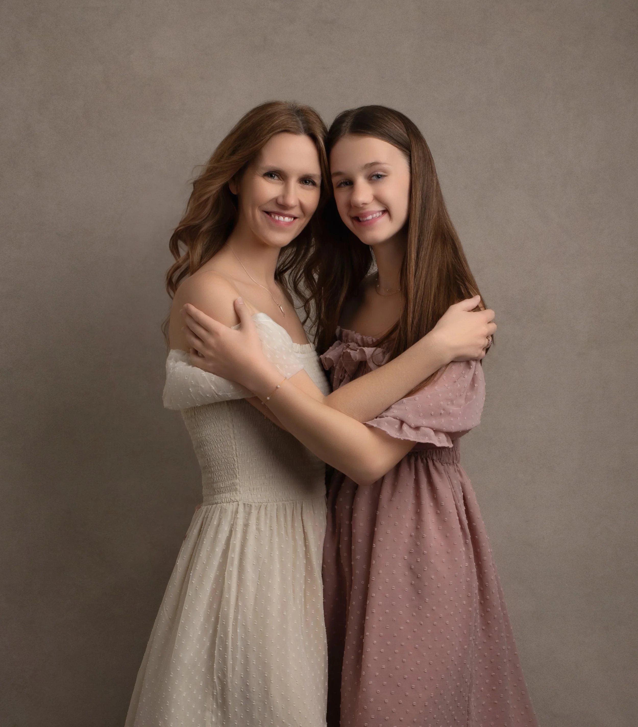 A woman and a young girl hug and smile at the camera against a neutral background.