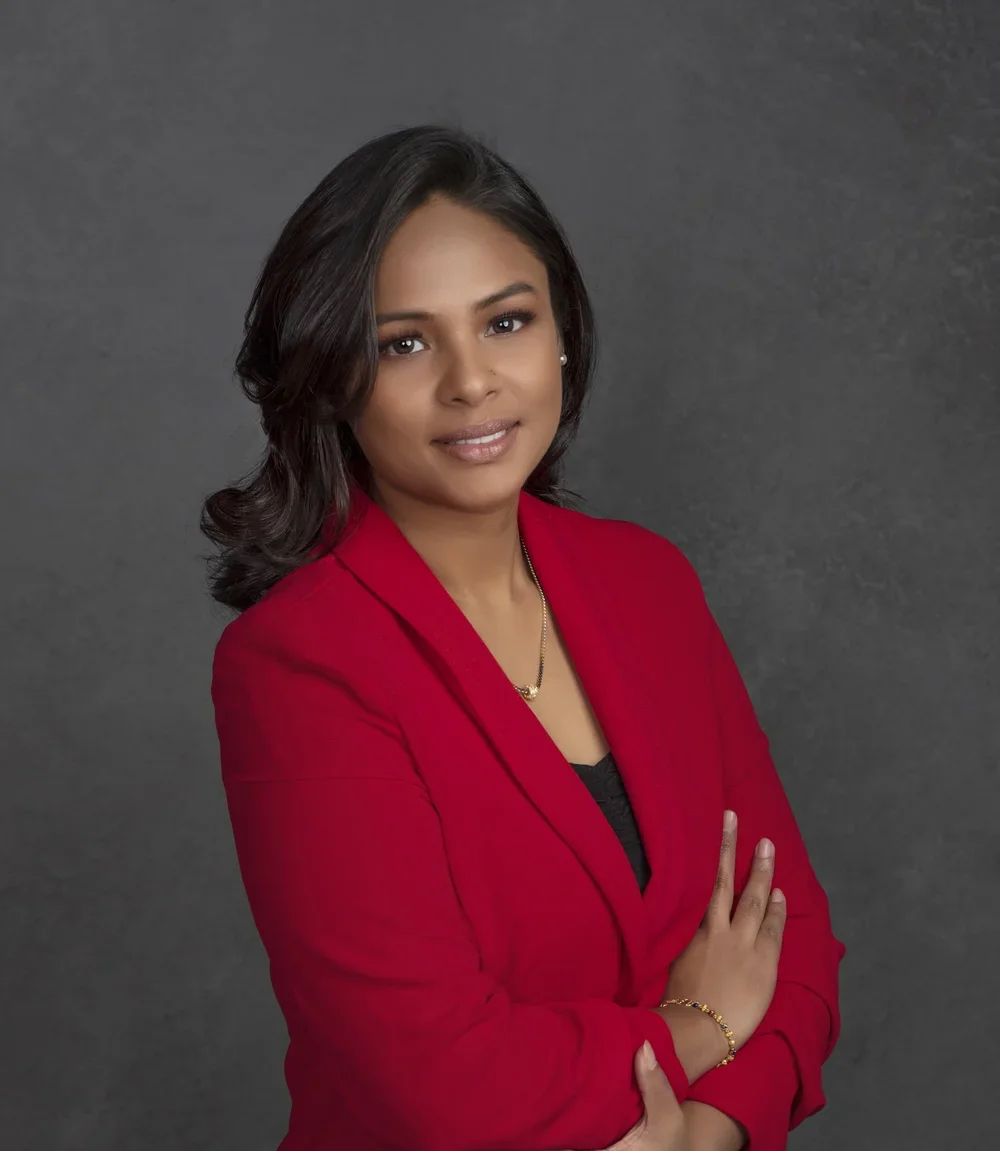 Photoshoot by Ally, A woman with shoulder-length dark hair, wearing a red blazer over a black top, standing with arms crossed against a gray background.