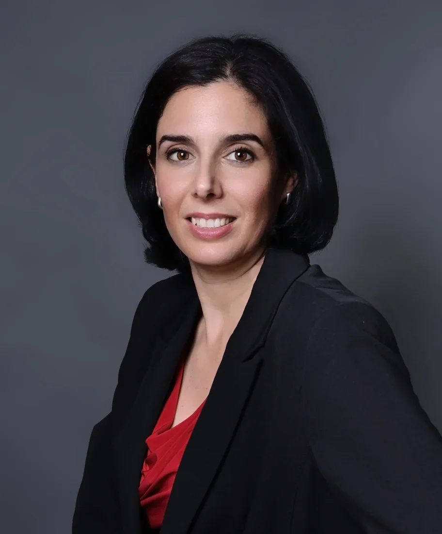 Photoshoot by Ally, A professional woman with dark hair, wearing a black blazer and a red blouse, smiling against a gray background.