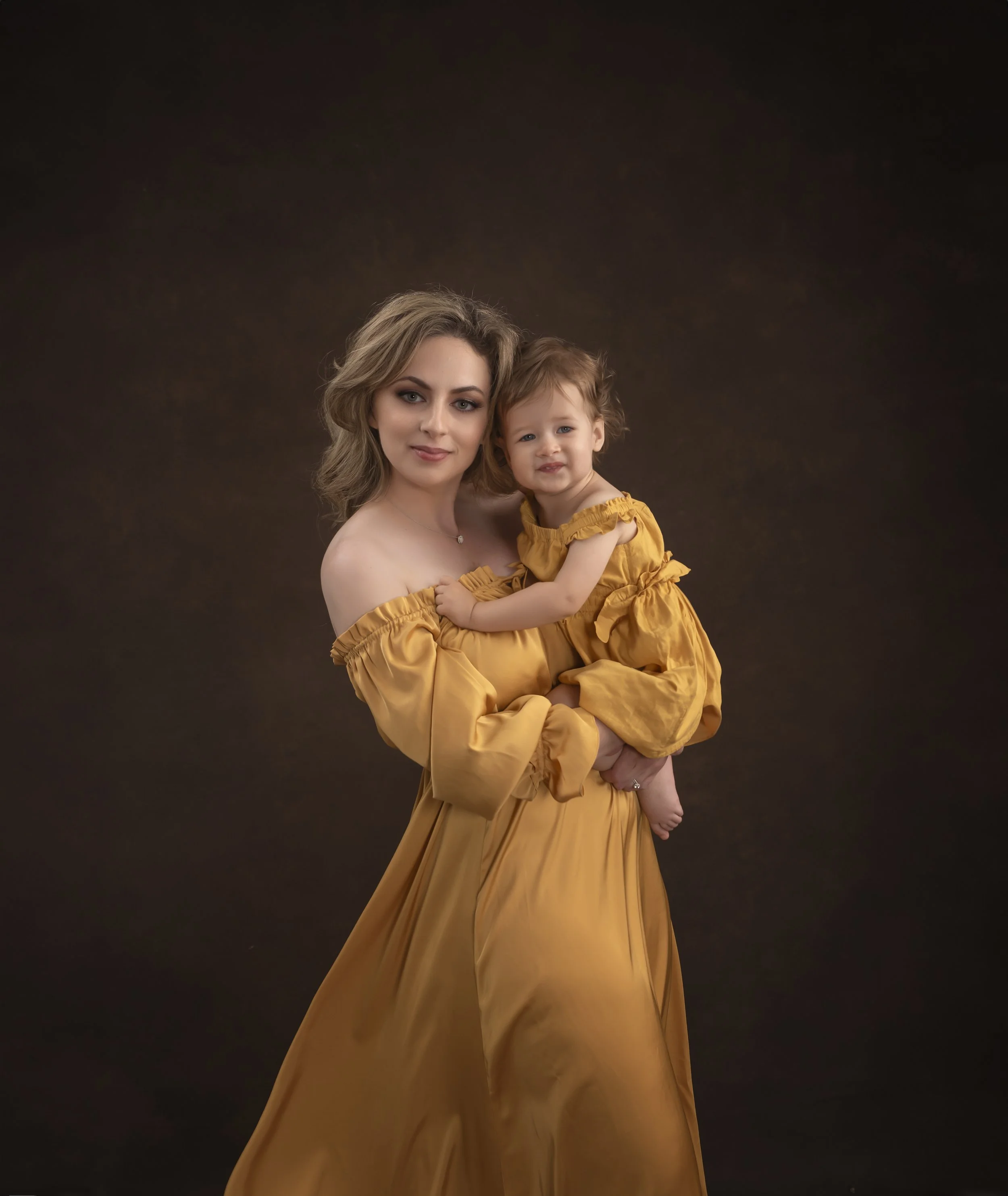 A woman in a mustard-yellow off-the-shoulder dress holds a young girl also dressed in a matching mustard-yellow dress, against a dark background.
