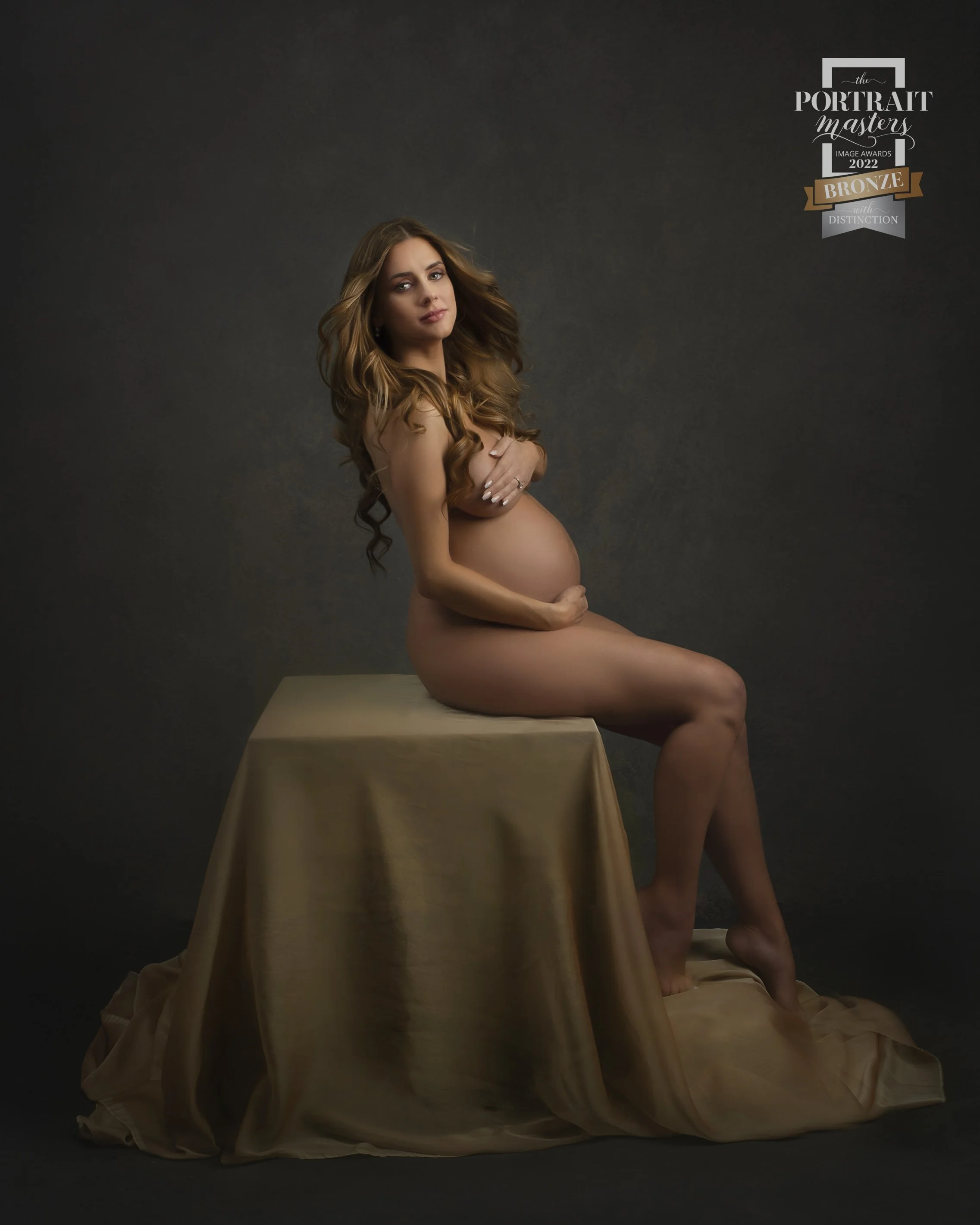 A pregnant woman with long, wavy hair sitting nude on a gold cloth-covered platform, holding her belly with one hand and resting her other hand on her chest, against a dark background. There is a label in the upper right corner indicating an award fo