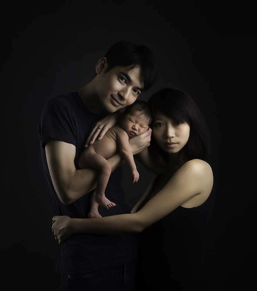 A young family with a man, woman, and newborn baby posing against a black background. The man holds the baby, and the woman supports the baby’s head, all looking at the camera.
