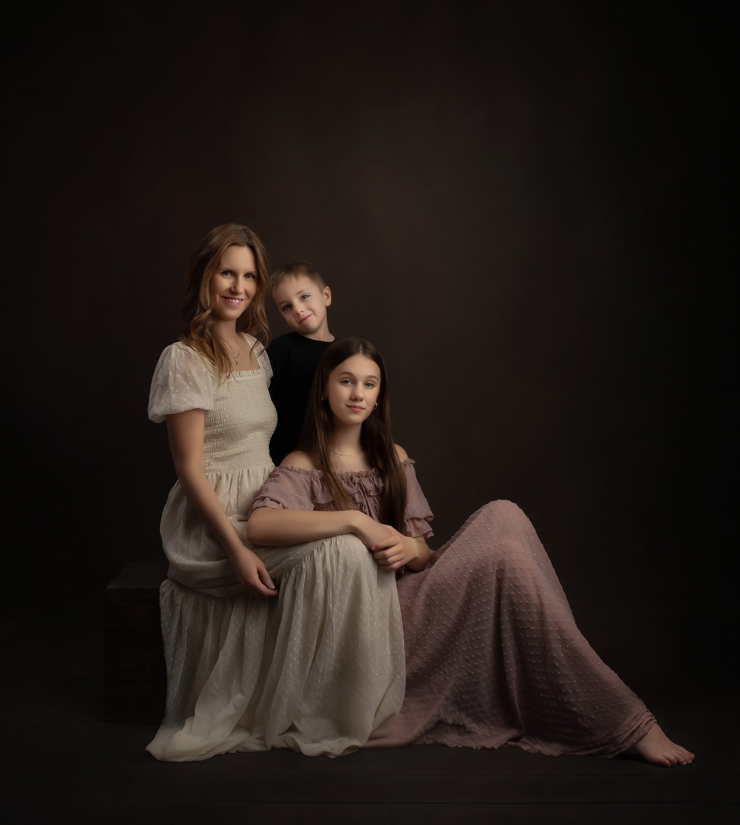 A woman and two children pose for a portrait against a dark background. The woman is standing on the left, wearing a cream-colored dress, smiling, with long wavy hair. The girl, seated on the right, wears a mauve off-the-shoulder dress with long brow