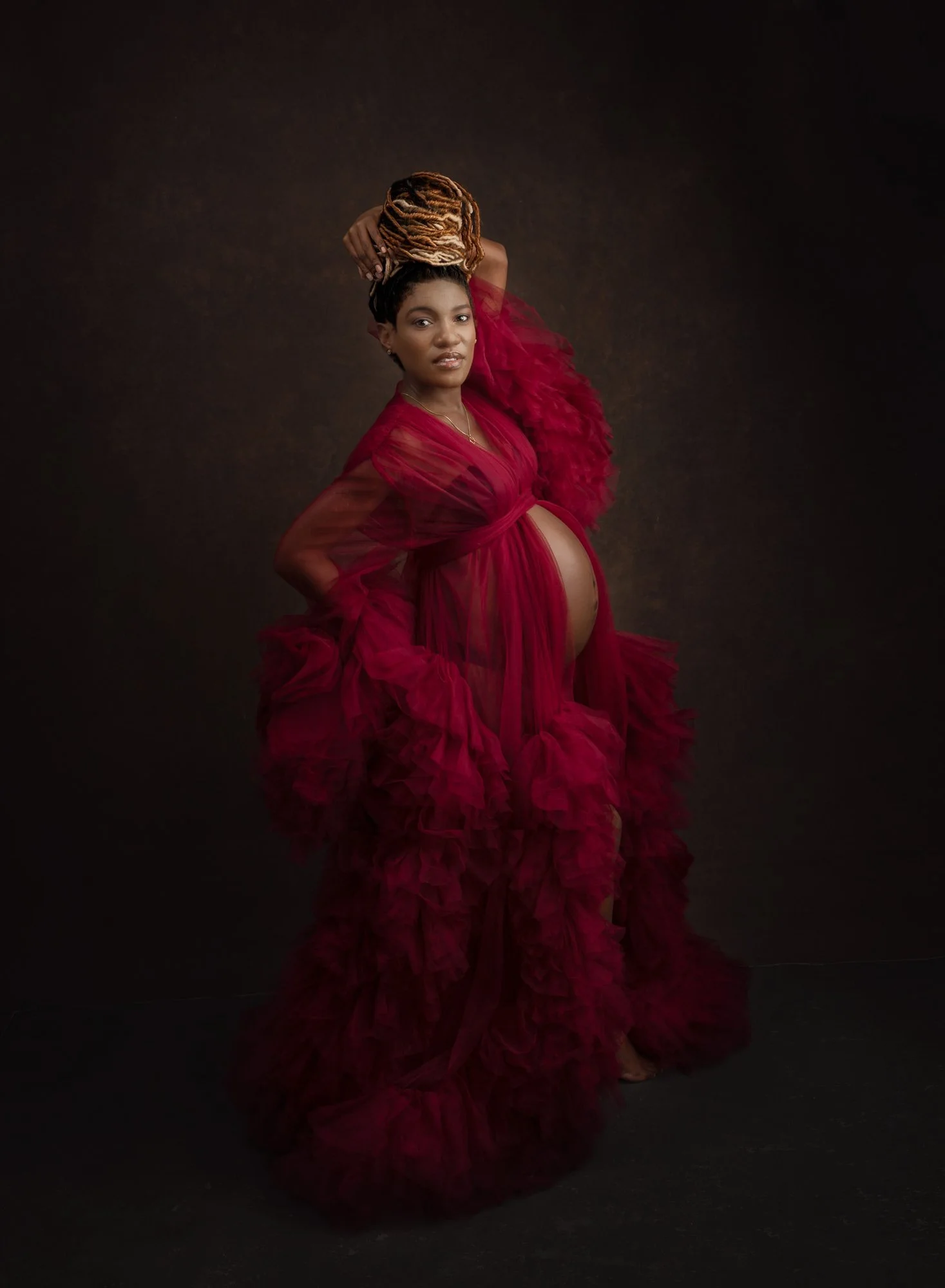 Pregnant woman in a flowing red dress posing with one arm raised, with a dark background.