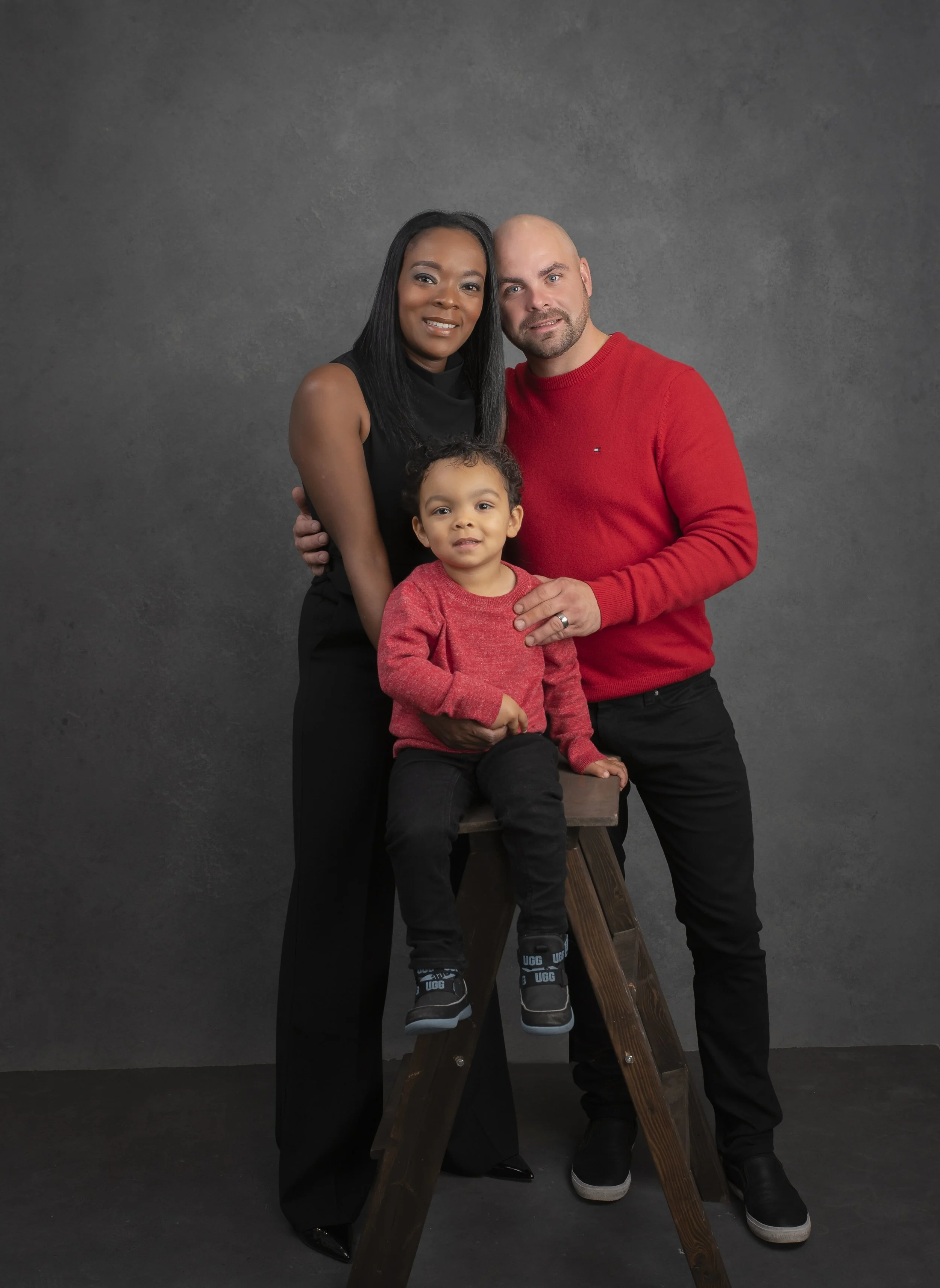 A family of three poses together against a dark gray background. The woman is standing on the left, wearing a sleeveless black dress, with long black hair and a warm smile. The man is on the right, wearing a red sweater and black pants, with a light 